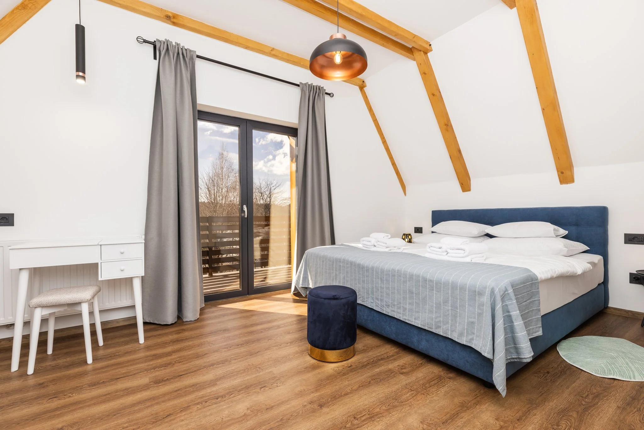 A bedroom with exposed wooden beams ceiling, a large window with gray curtains, a bed with a light blue bedspread, a navy blue ottoman, a small white desk with a stool, and wooden flooring.