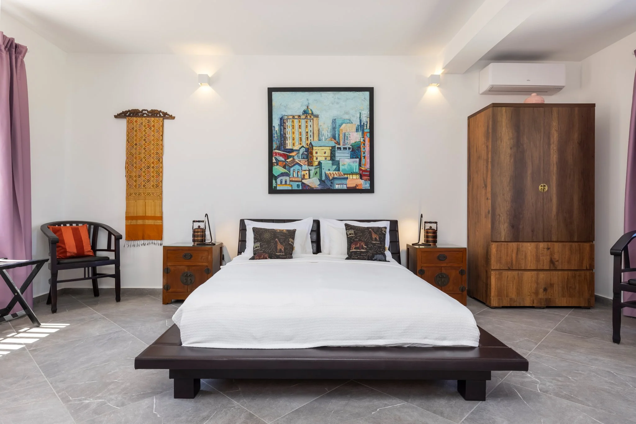 A neatly made bed with white bedding and decorative pillows featuring animal motifs in a modern bedroom. The room is decorated with a framed cityscape painting above the bed, wooden bedside tables with lamps, purple curtains, a wooden wardrobe, and b