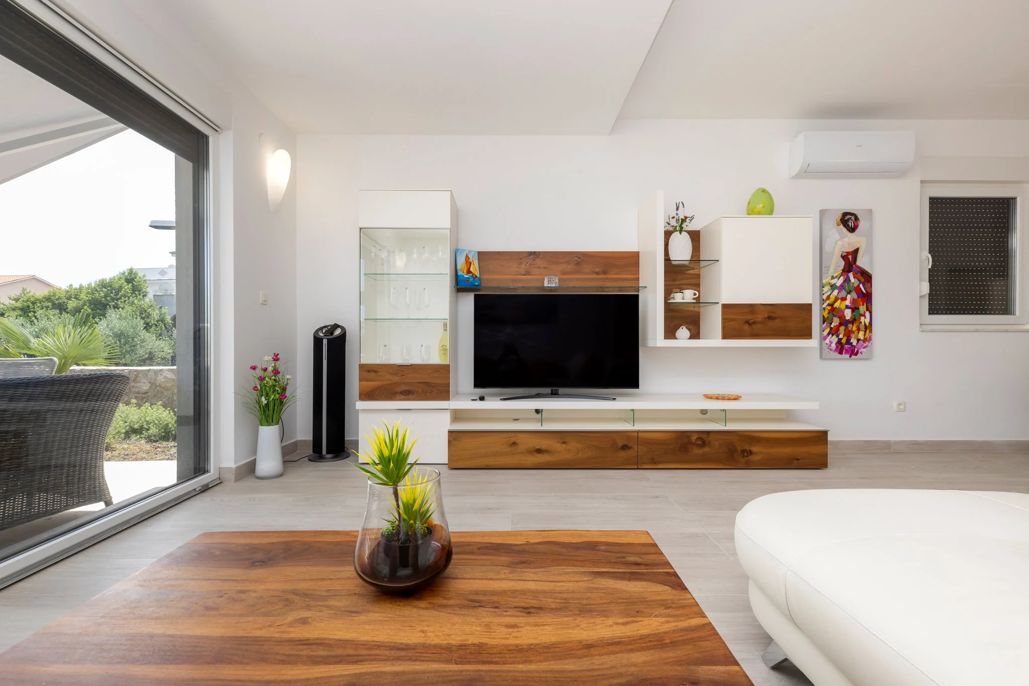 A modern living room with white walls and furniture, a flat-screen TV on a wooden and white entertainment unit, two glass shelves, and various decorative items including a painting, vases, and plants; a sliding glass door opens to a patio with outdoo