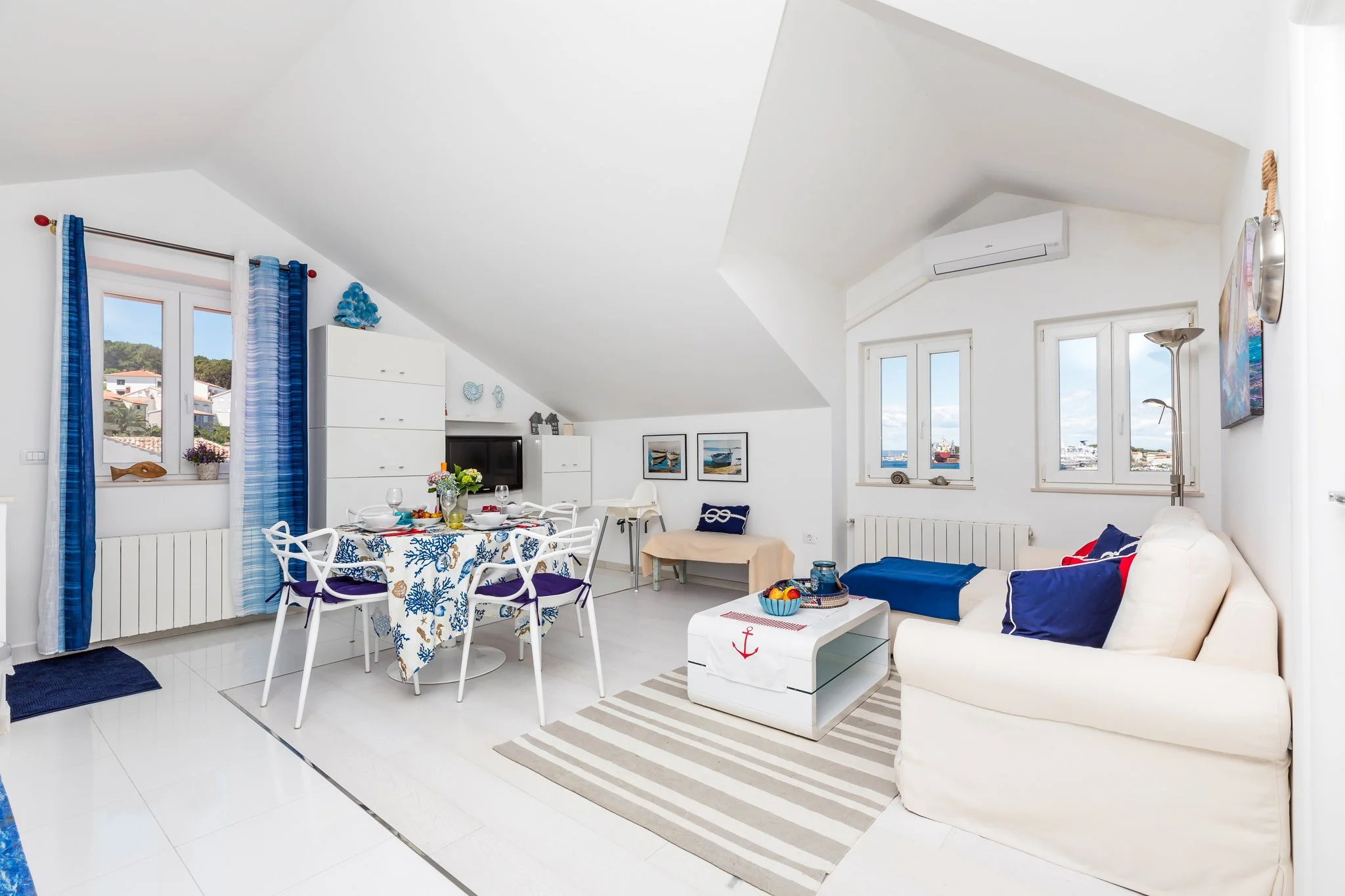 Interior of a bright and airy living room with white walls and ceiling. Features include a dining table with six white chairs, a white sofa with blue and red cushions, and large windows overlooking a cityscape. Decorative items include nautical-theme