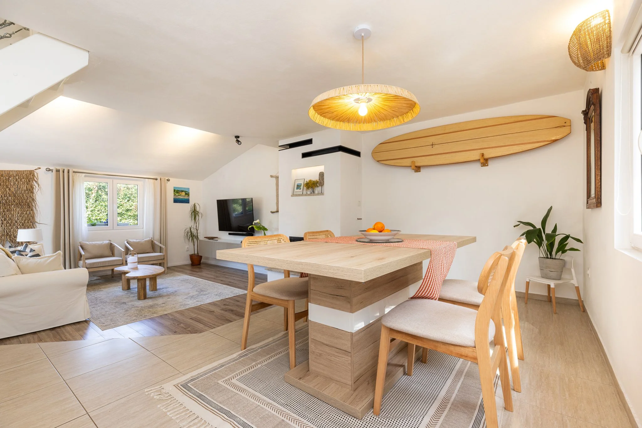 Living room and dining area with a wooden dining table, light wood chairs, a white couch, armchairs, a wall-mounted TV, plants, large windows with curtains, and surfboards on the wall.
