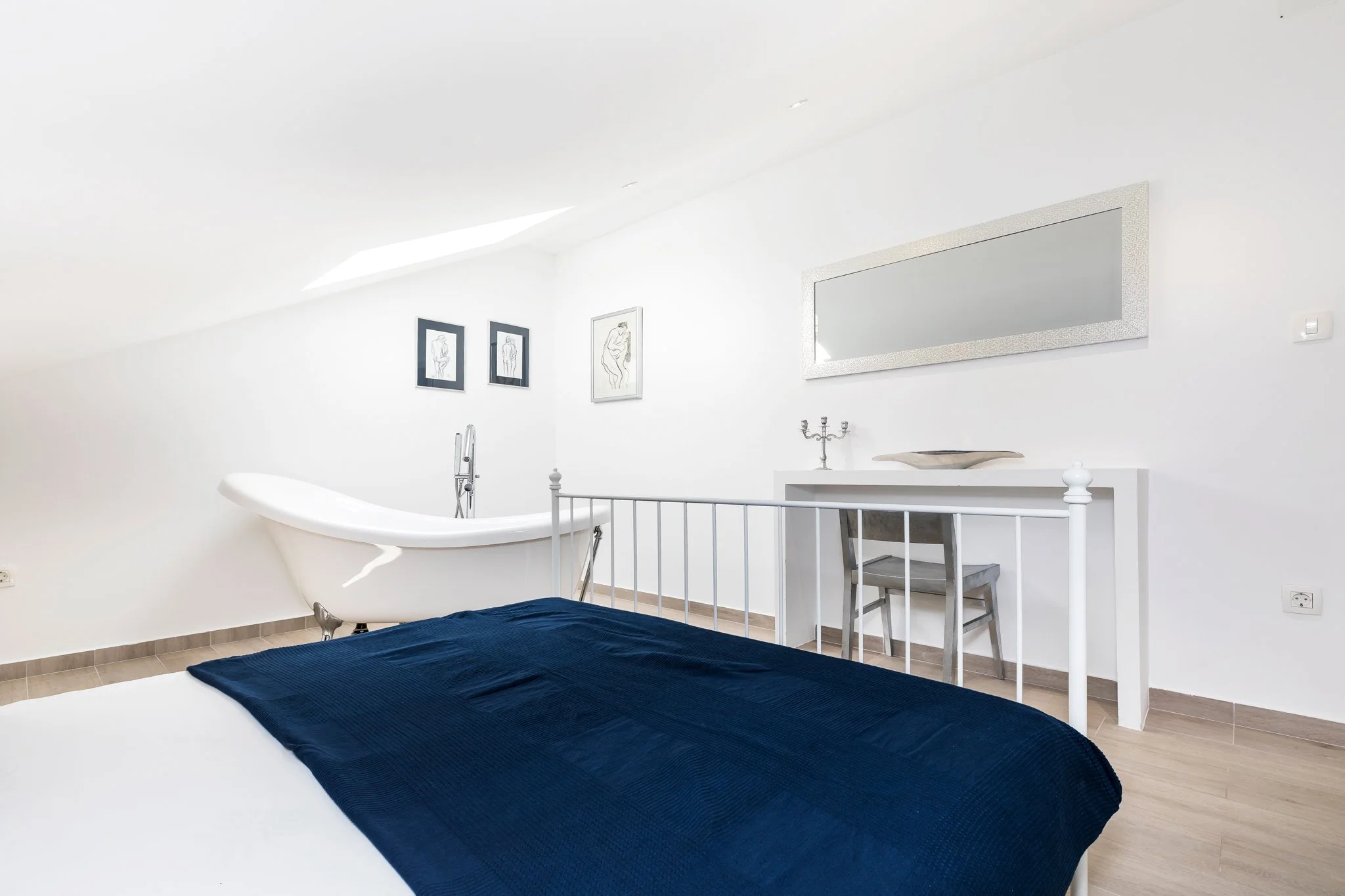 A bedroom with a white metal bed frame, a dark blue bedspread, and a white dresser with a mirror and artwork on the wall.