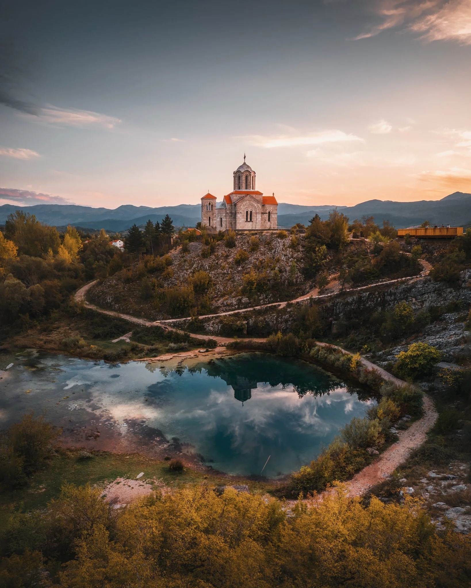 A church on a hill surrounded by trees, with mountains and a lake in the background during sunset.