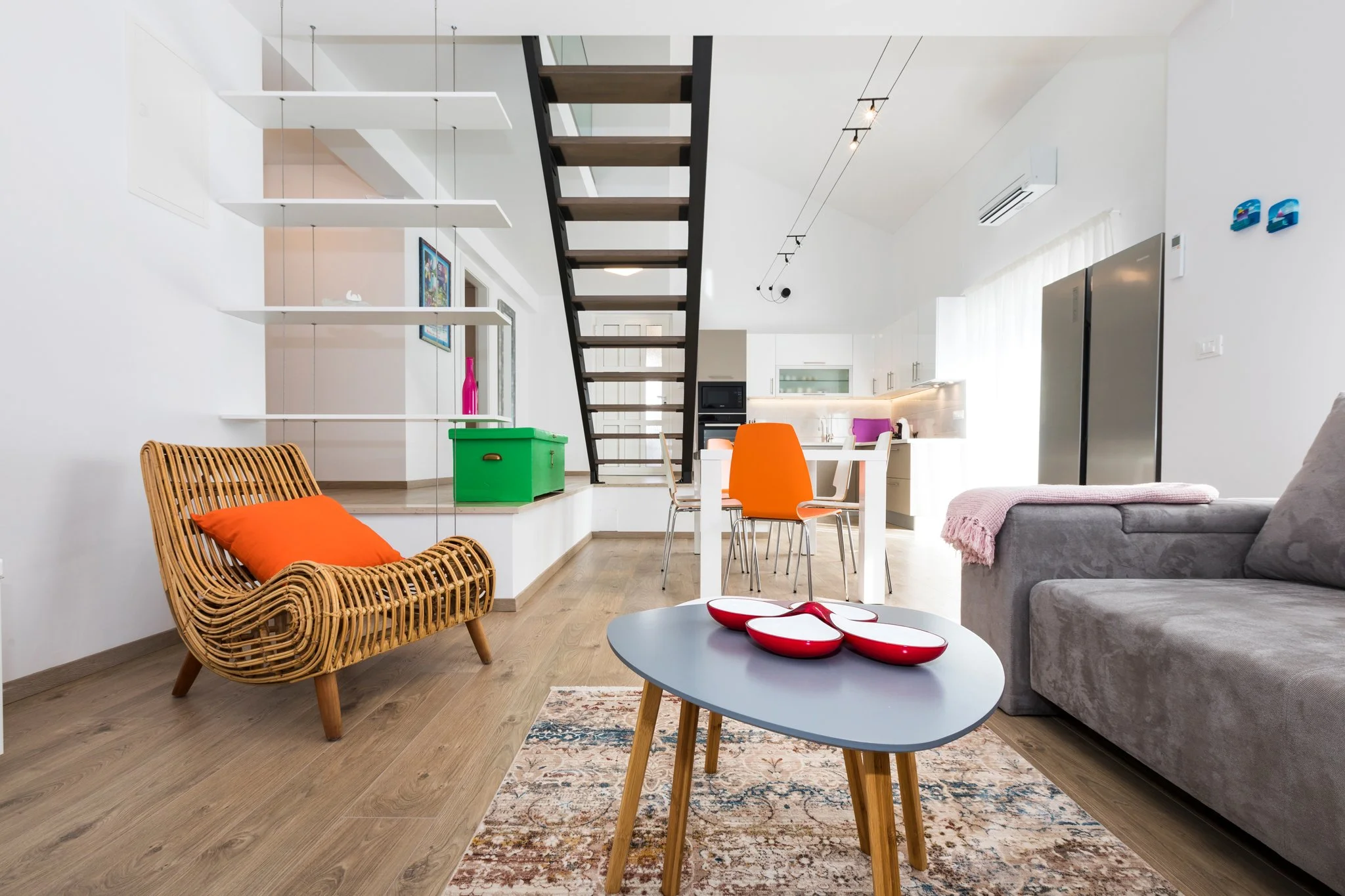 Interior of a modern, open-plan living space with white walls and wooden flooring. Features include a rattan lounge chair with an orange cushion, a gray sofa with a pink throw, a white dining table with white and orange chairs, a coffee table with re