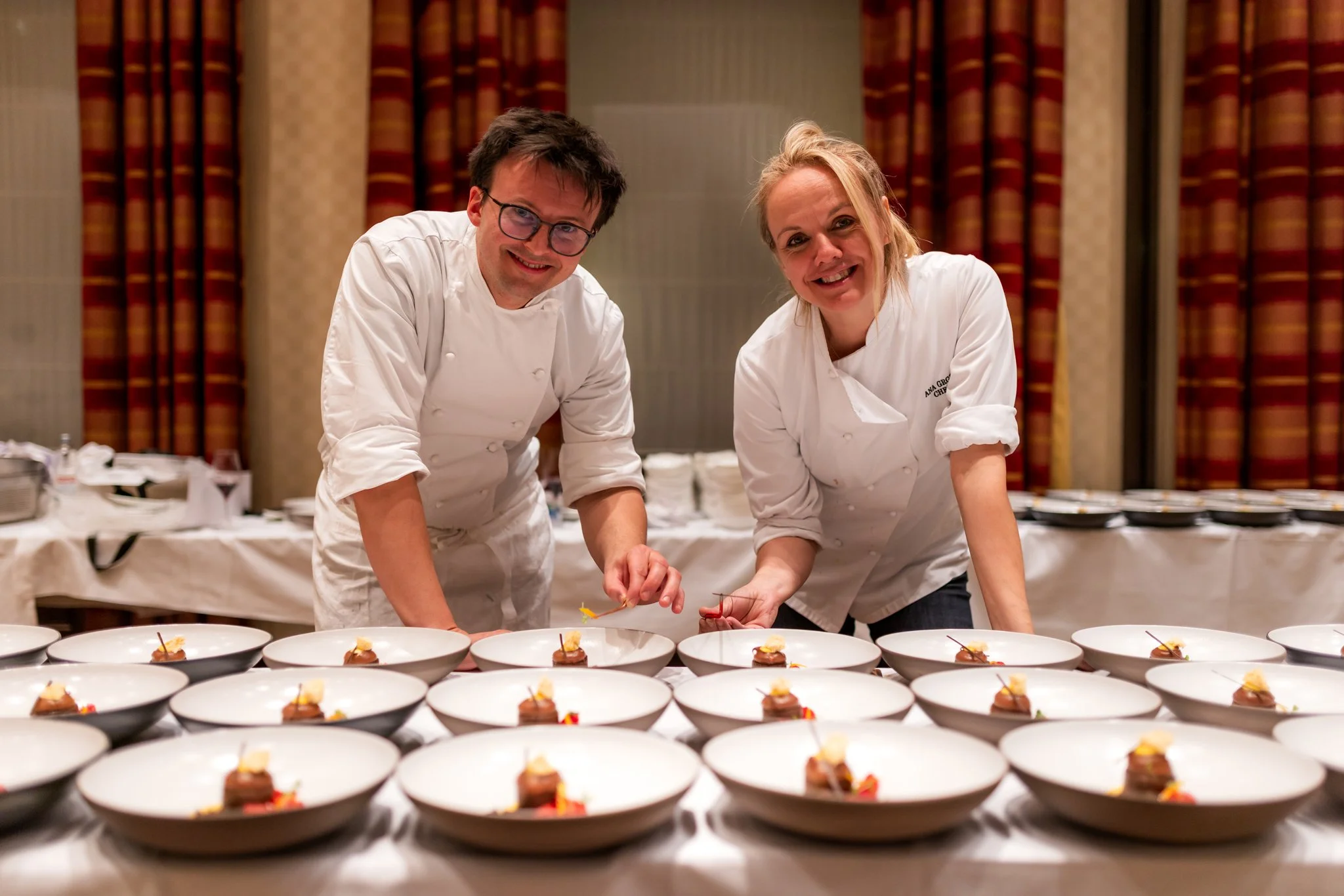 Two chefs in white uniforms are preparing plated dishes in a professional kitchen or banquet setting, with numerous identical dishes arranged on a long table in front of them.