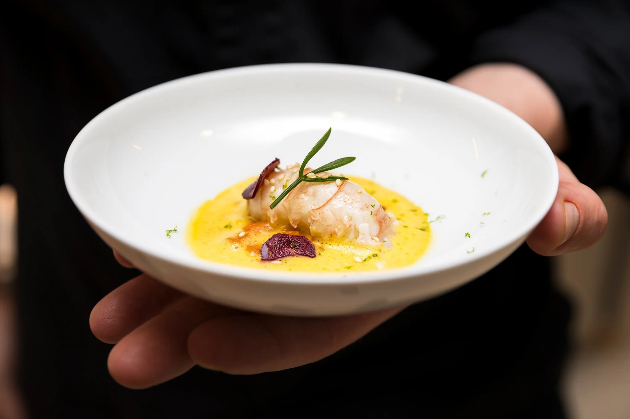 A person holding a white bowl with a yellow sauce, topped with a piece of seafood, garnished with green herbs, sesame seeds, and red vegetable pieces.