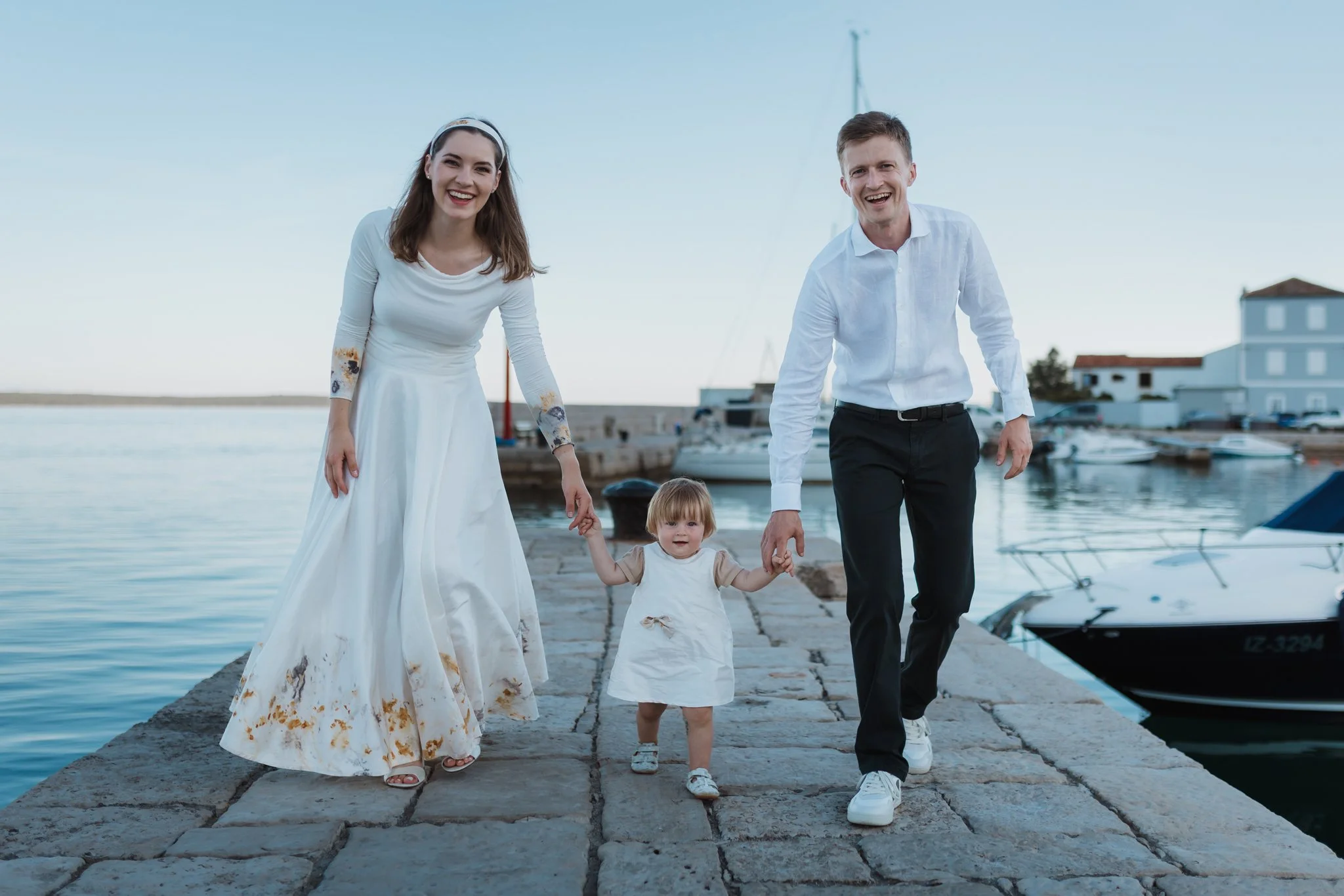 A family of four walking hand in hand on a dock near boats and water, smiling and enjoying a day outdoors.