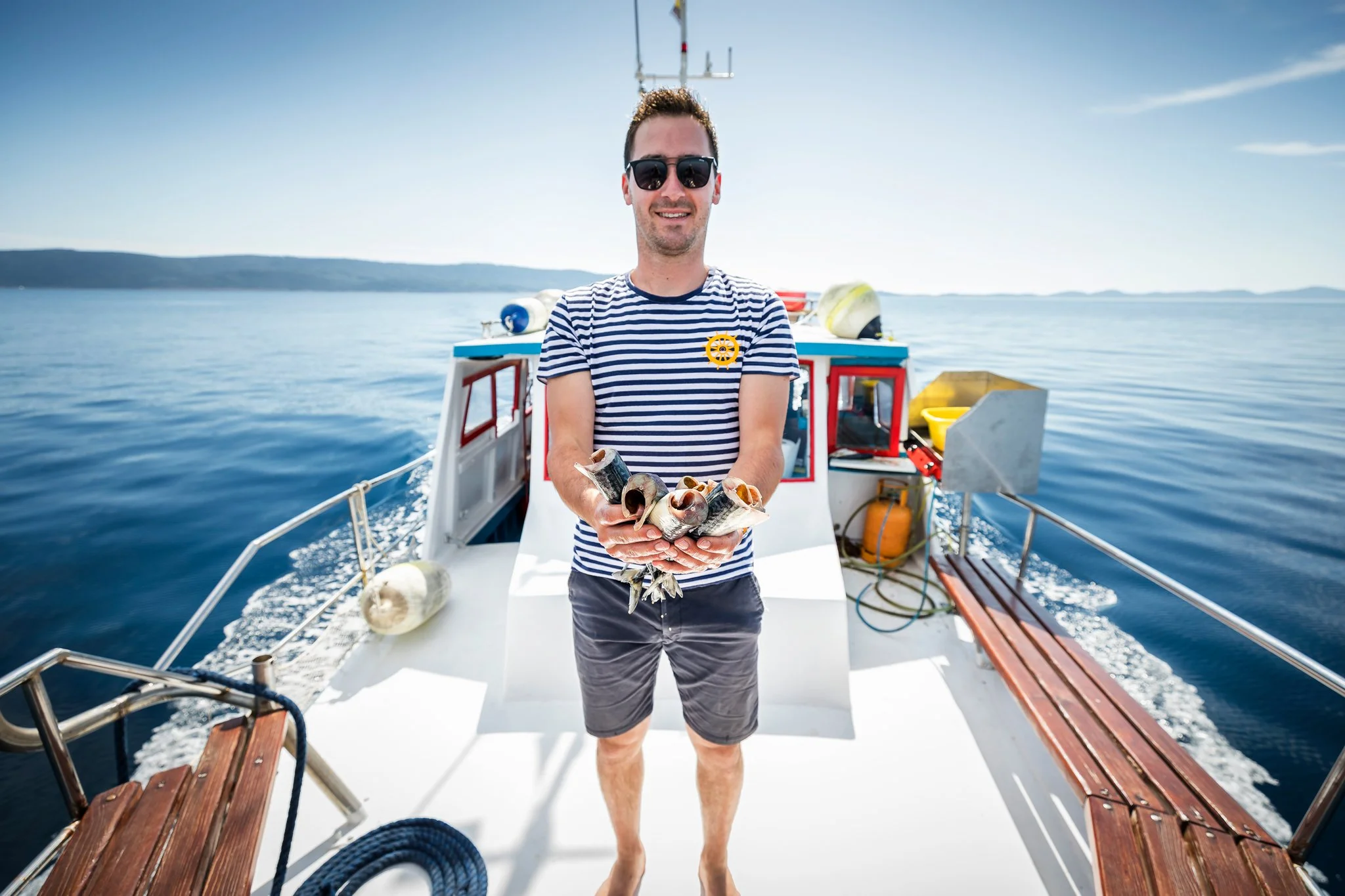 A man wearing sunglasses, a striped shirt with a yellow badge, and shorts standing on the deck of a boat holding seashells, with water and a distant shoreline in the background on a sunny day.