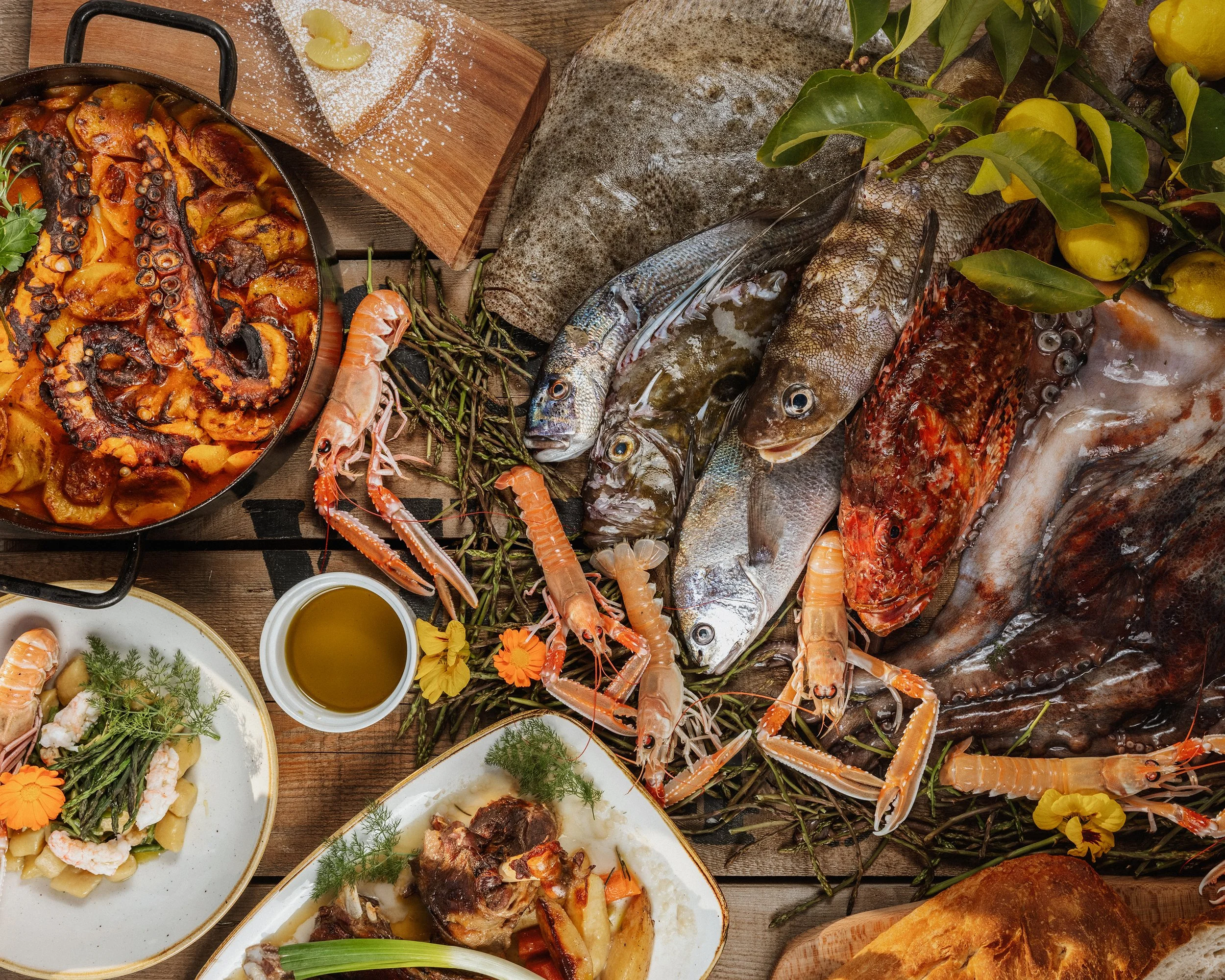 An assortment of seafood dishes on a rustic wooden table, including fish, shrimp, and shellfish, alongside a plate of pasta salad and a baked casserole, with lemon wedges, green herbs, and small bowls of sauce or oil.