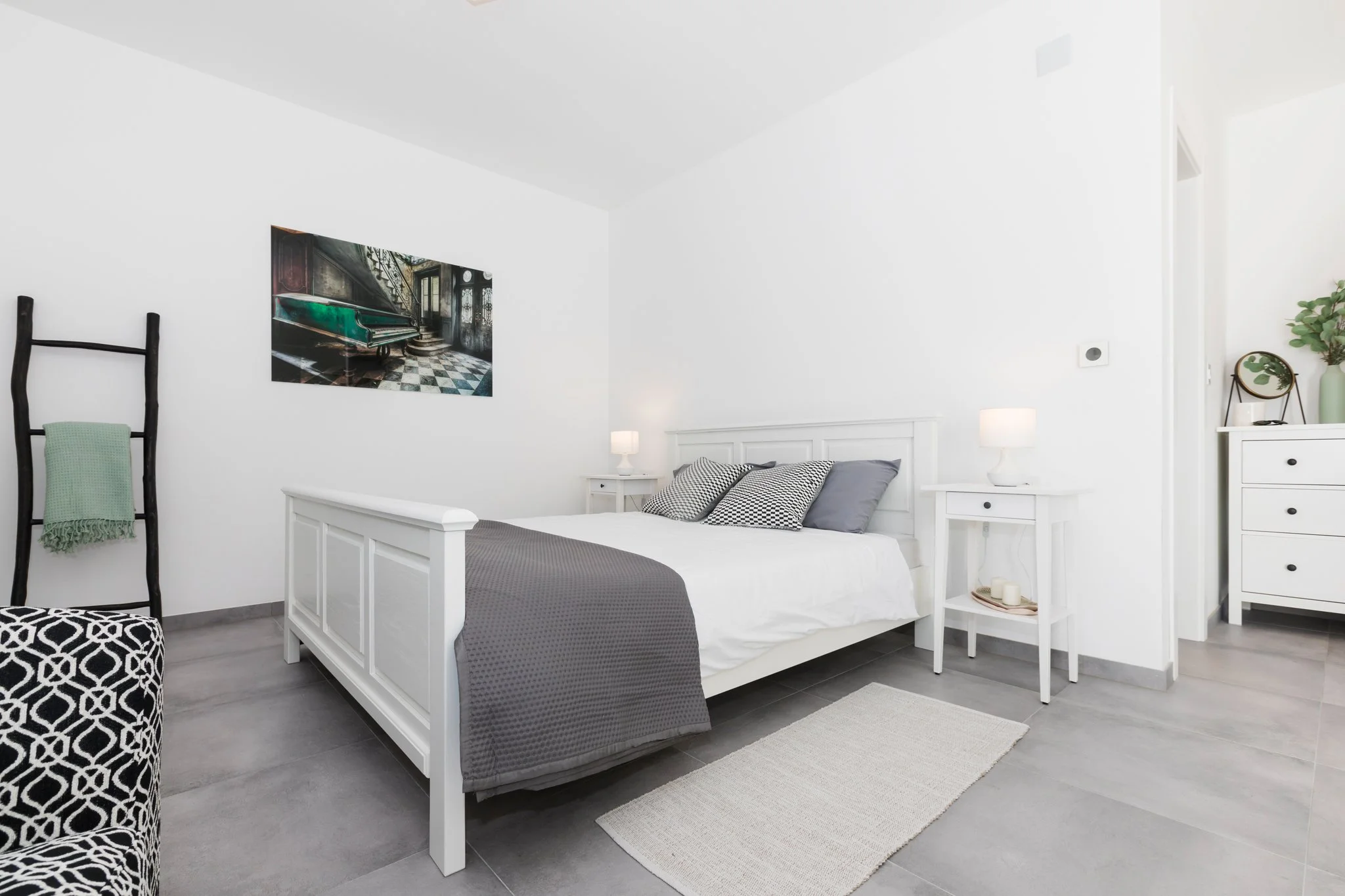 A minimalist bedroom with white furniture, a white bed with gray and black patterned pillows, black and white patterned ottoman, painting of an interior with stairs, a green vase with a plant, and black ladder with a green towel.