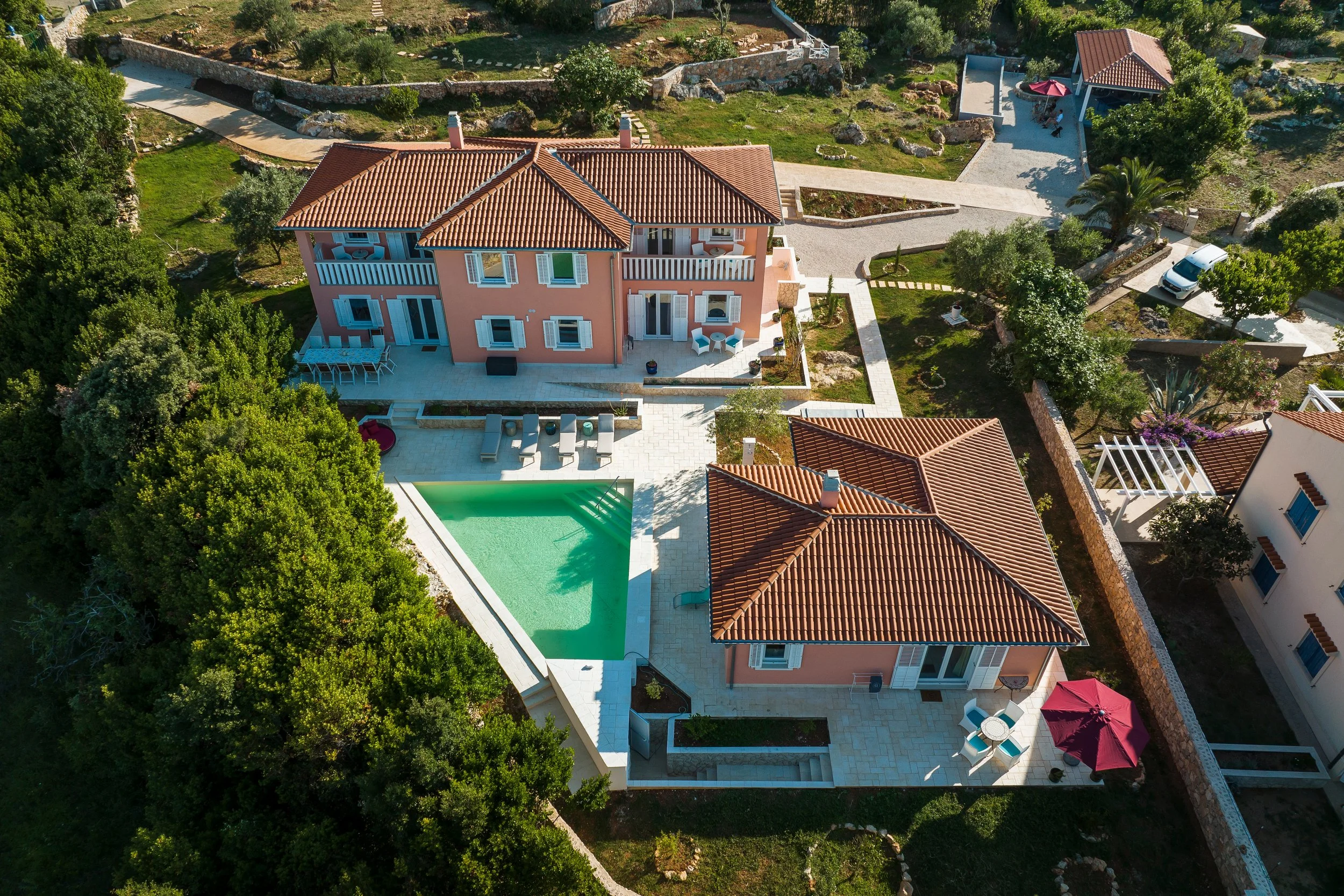 Aerial view of a pink two-story house with a red-tiled roof, a swimming pool, outdoor seating areas with umbrellas, surrounded by greenery and neighboring houses with similar roofs.