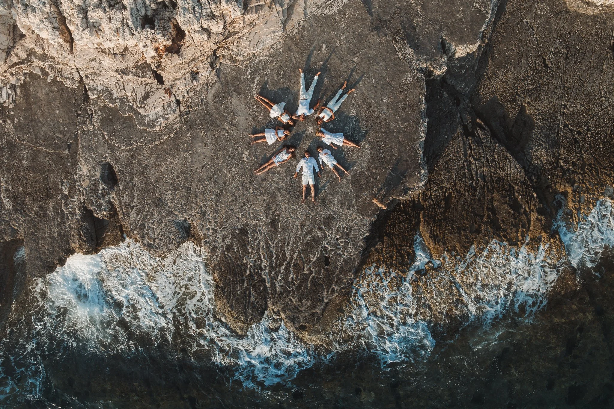 Six women lying on a rocky surface near the ocean, arranged in a star-like pattern, wearing white clothing. The photo is taken from above at sunset.