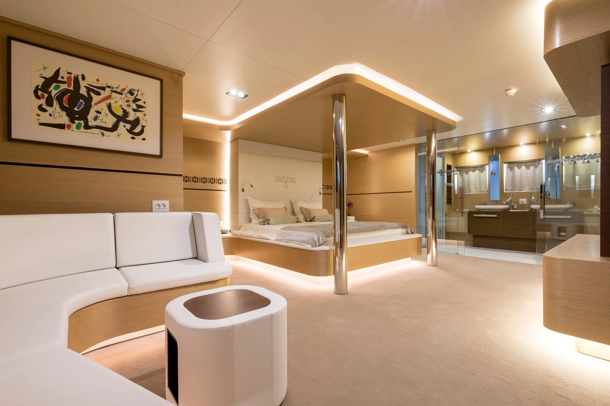 Modern hotel room with a curved white sofa, wooden accents, a bed in the background, and a glass-enclosed bathroom area with sinks.