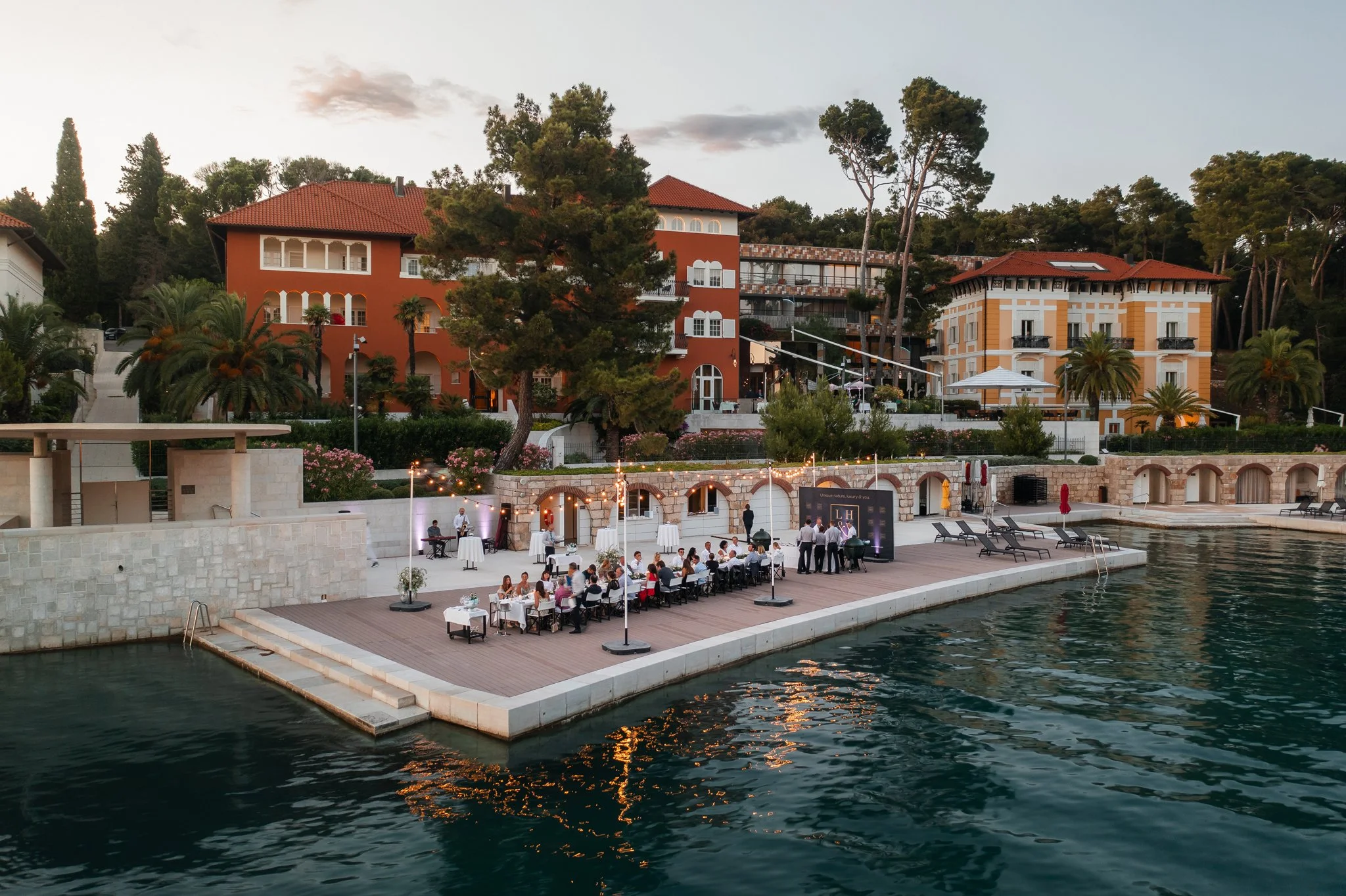 An elegant outdoor waterfront dining area at sunset with tables, chairs, and string lights, adjacent to colorful residential buildings with red-tiled roofs, surrounded by lush trees.Alfred Keller in Mali Lošinj, Boutique Hotel Alhambra.