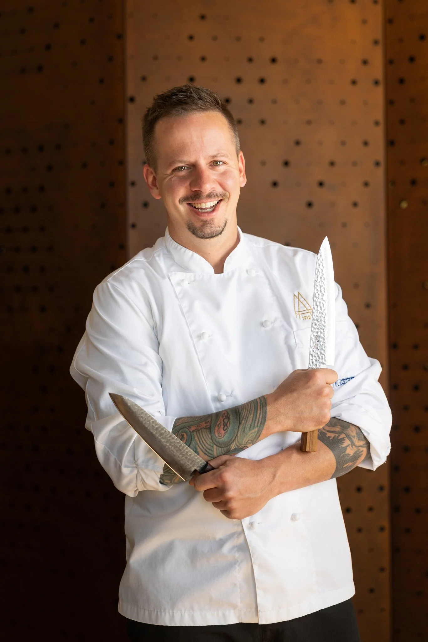 A smiling male chef with tattoos wearing a white chef's coat, holding a large kitchen knife, standing in front of a wooden background with perforations.