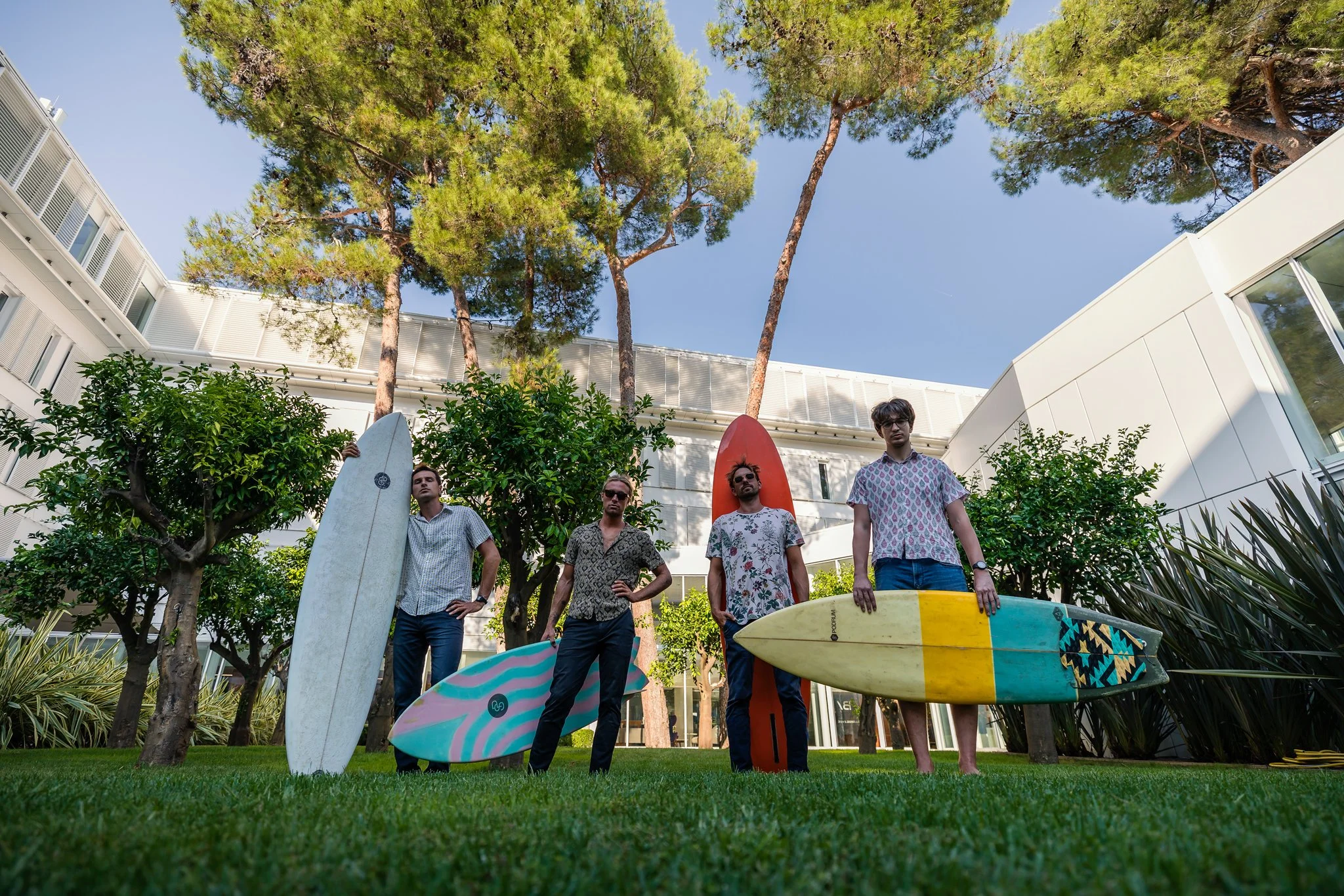 Four young men standing outdoors in a grassy area with trees and modern buildings behind them, each holding or standing near colorful surfboards.