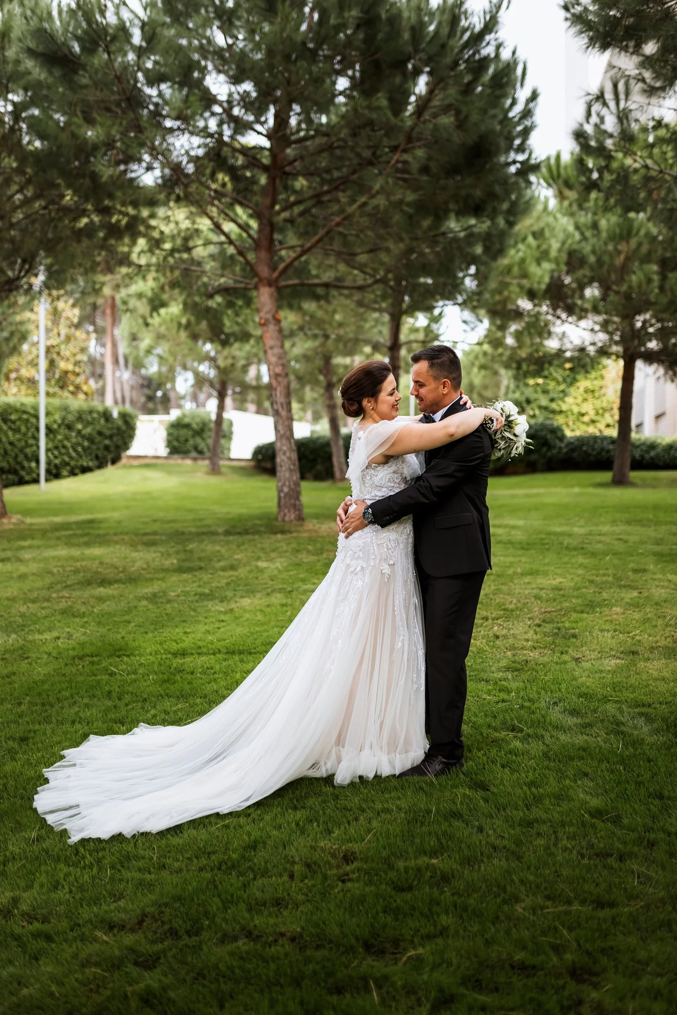 A bride and groom embrace outdoors in a park with green grass and tall trees in the background. The bride wears a white lace wedding dress with a long train and holds a bouquet, smiling at the groom. The groom is dressed in a black suit and tie, smil