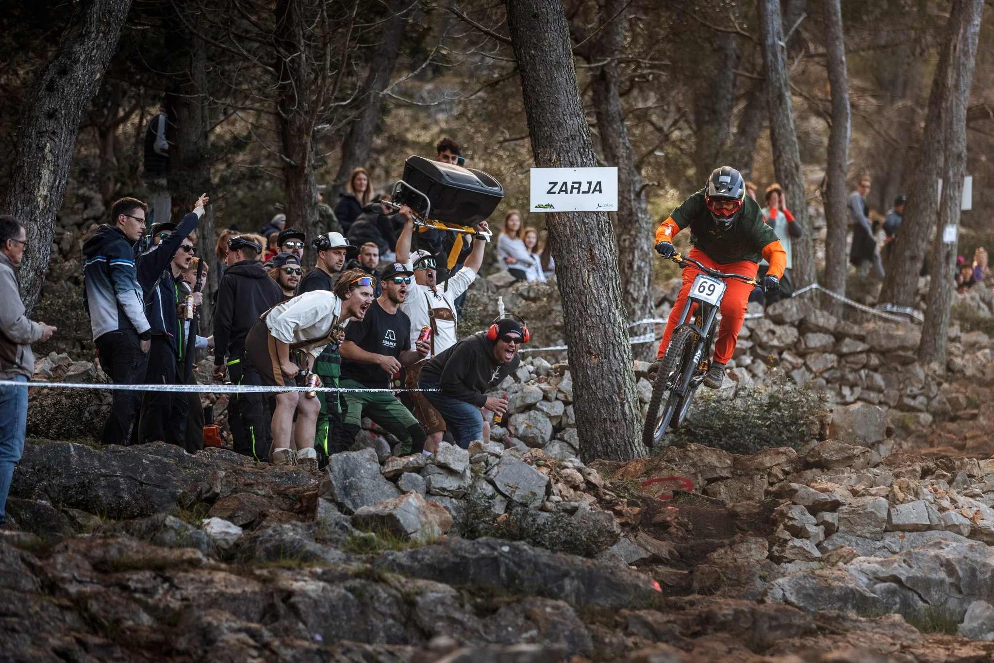 A mountain biker wearing a helmet and orange and black gear is riding downhill on a rocky terrain through a forest. There is a crowd of spectators on the side of the trail, some cheering, and some taking photos. One person is holding a sign that read