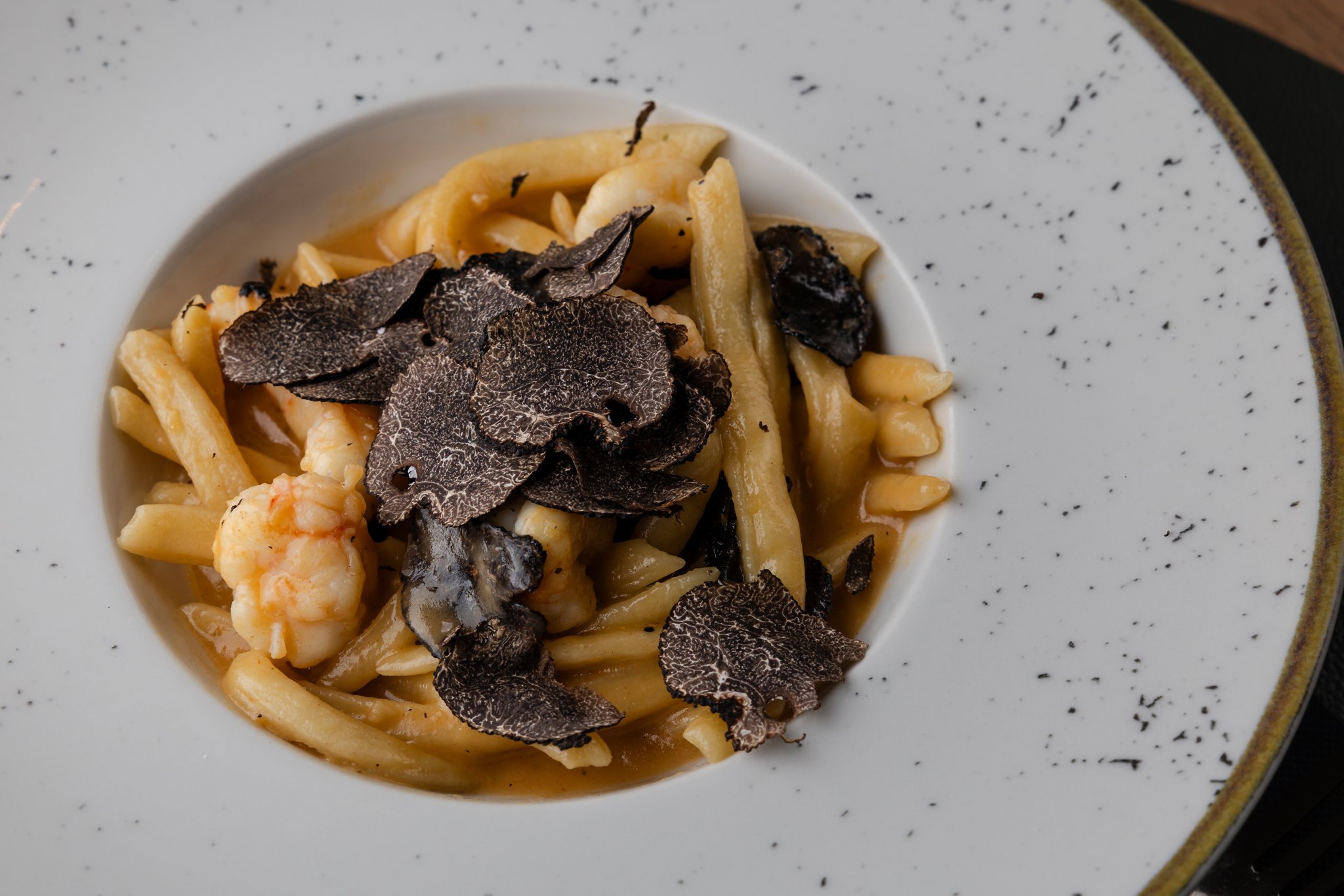 A plate of pasta with black truffle shavings on top, garnished with shrimp, served on a speckled white ceramic plate.