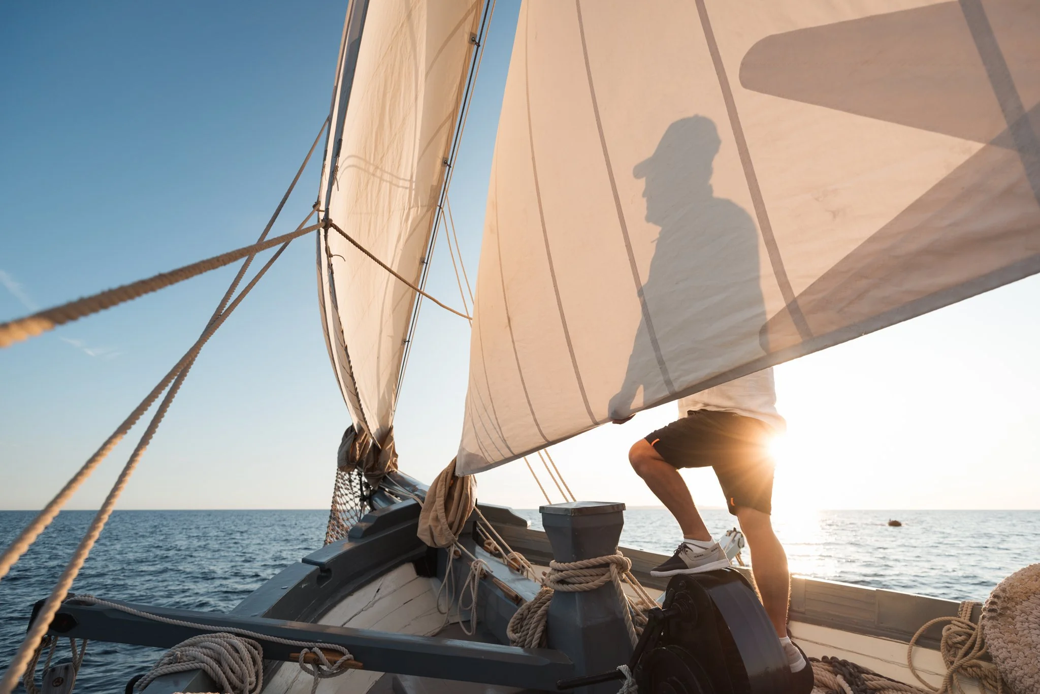 A person operating a sailboat on the water during sunset, with the shadow of their head cast on the sail, wearing shorts and sneakers, and surrounded by ropes and boat equipment.