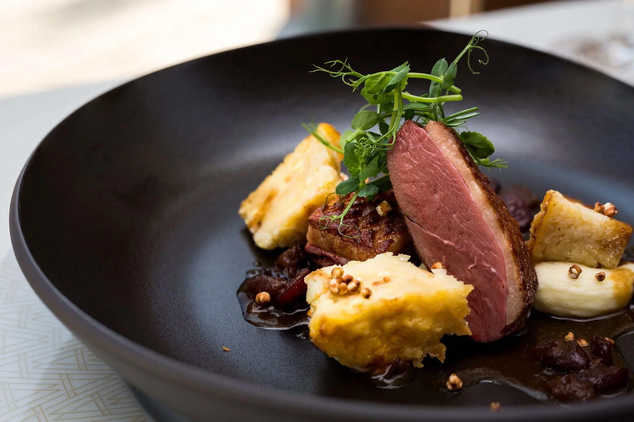 Close-up of a plated gourmet dish featuring a medium-rare cut of beef, roasted potatoes, greens, and garnishes in a black bowl.