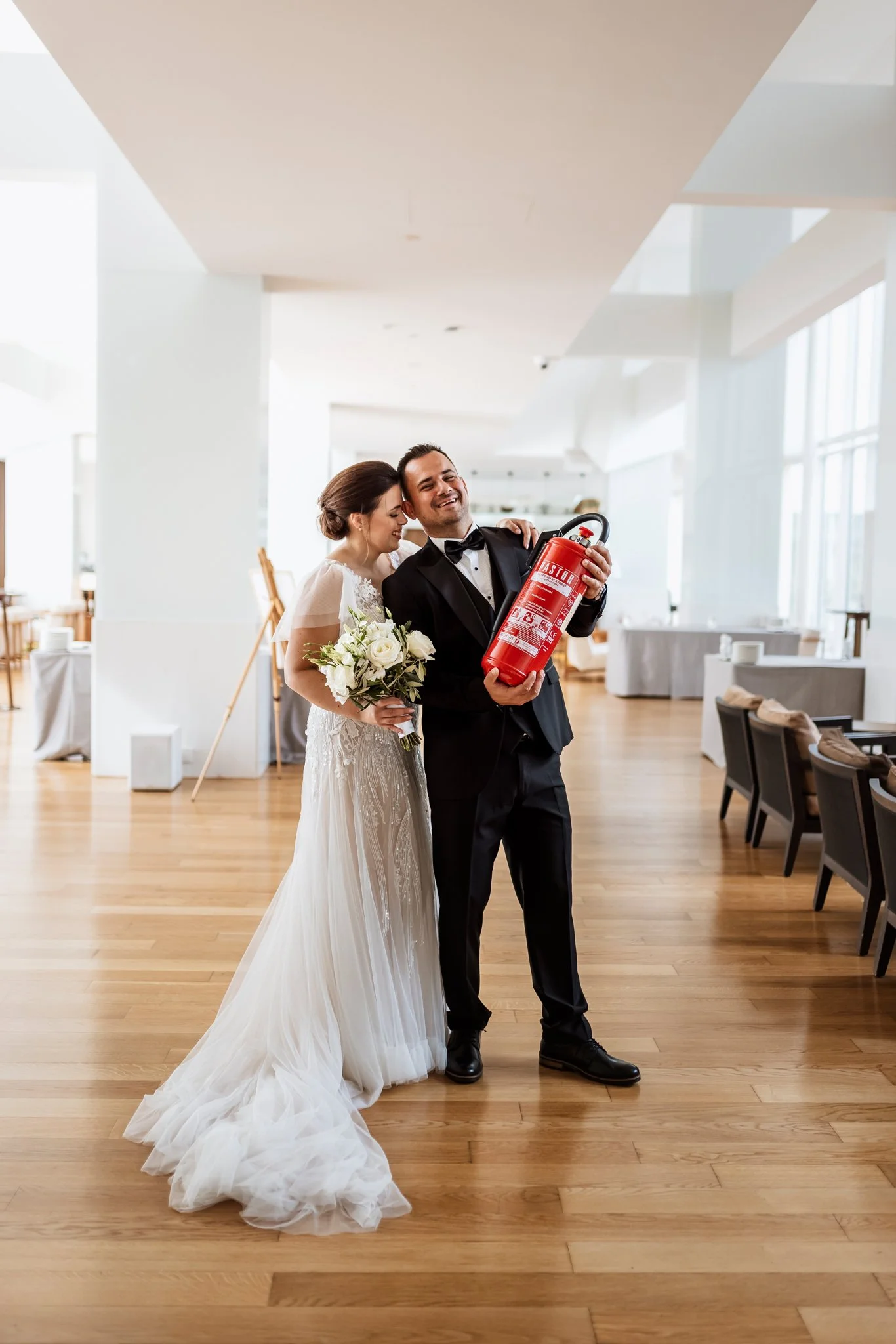 A bride and groom smiling at each other in a bright, modern wedding venue. The groom holds a fire extinguisher. The bride is holding a bouquet of white roses.