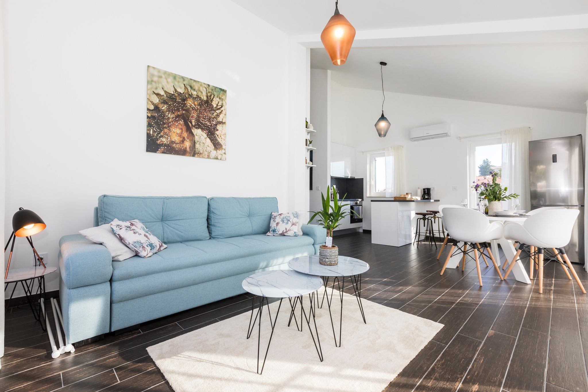 A modern living room with a blue sofa, two round marble coffee tables, a white rug, potted plants, and wall art of a sea creature. There's a contemporary kitchen and dining area in the background with hanging pendant lights, a window with curtains, a