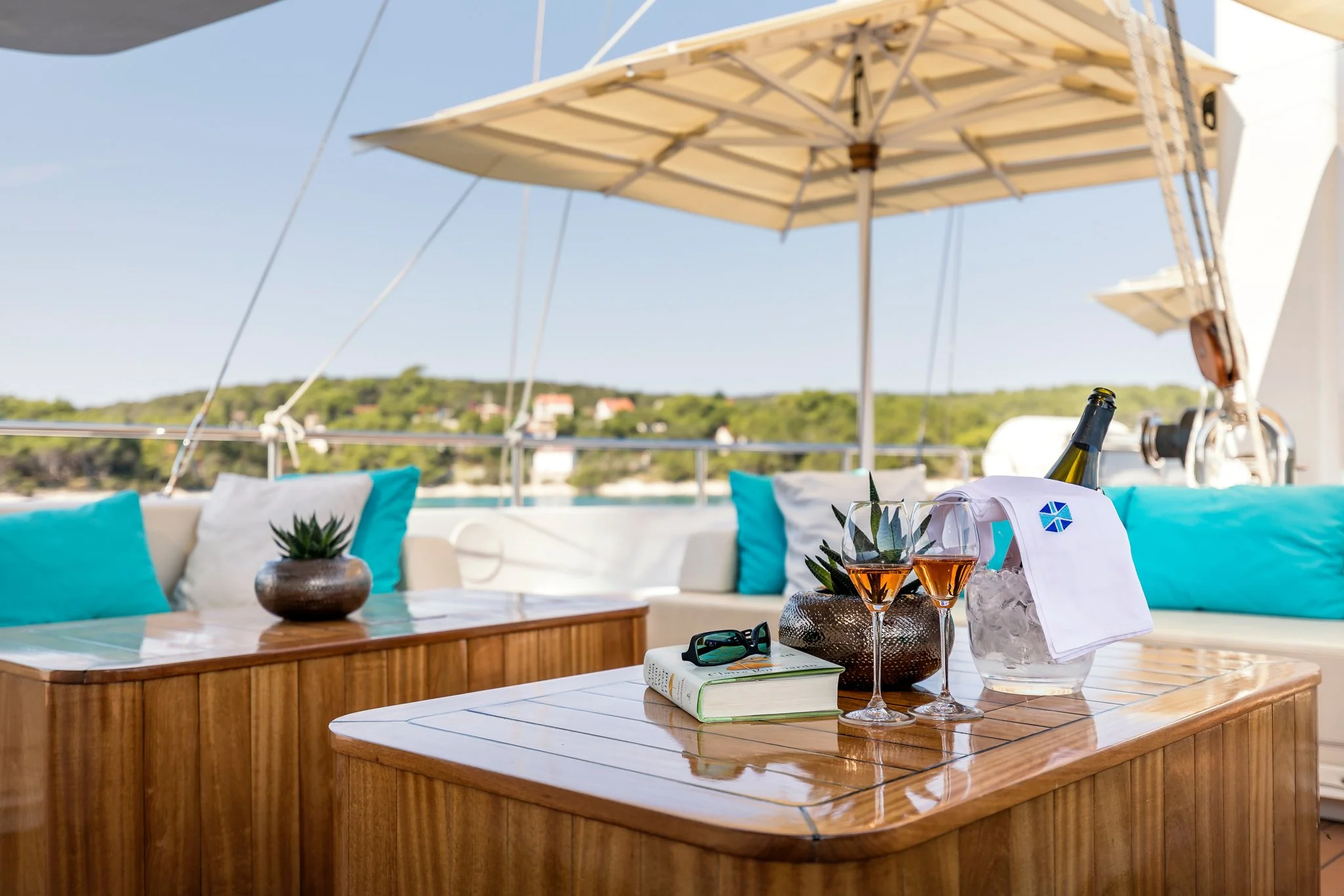 A wooden outdoor table with glasses of rosé wine, a small potted succulent, a wine bottle, and sunglasses, set against a background of a sofa with turquoise and white pillows, and large umbrellas on a balcony overlooking a green landscape.