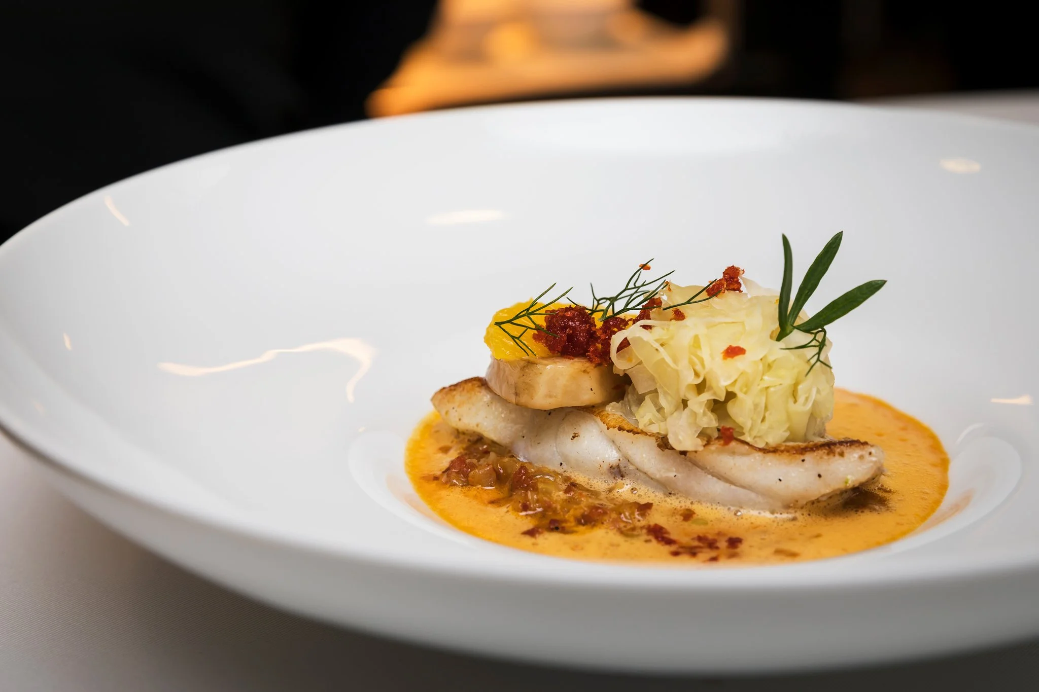 A plated dish featuring a piece of cooked fish topped with diced yellow vegetables, garnished with green herbs and small red crumbles, served with a flavorful sauce.