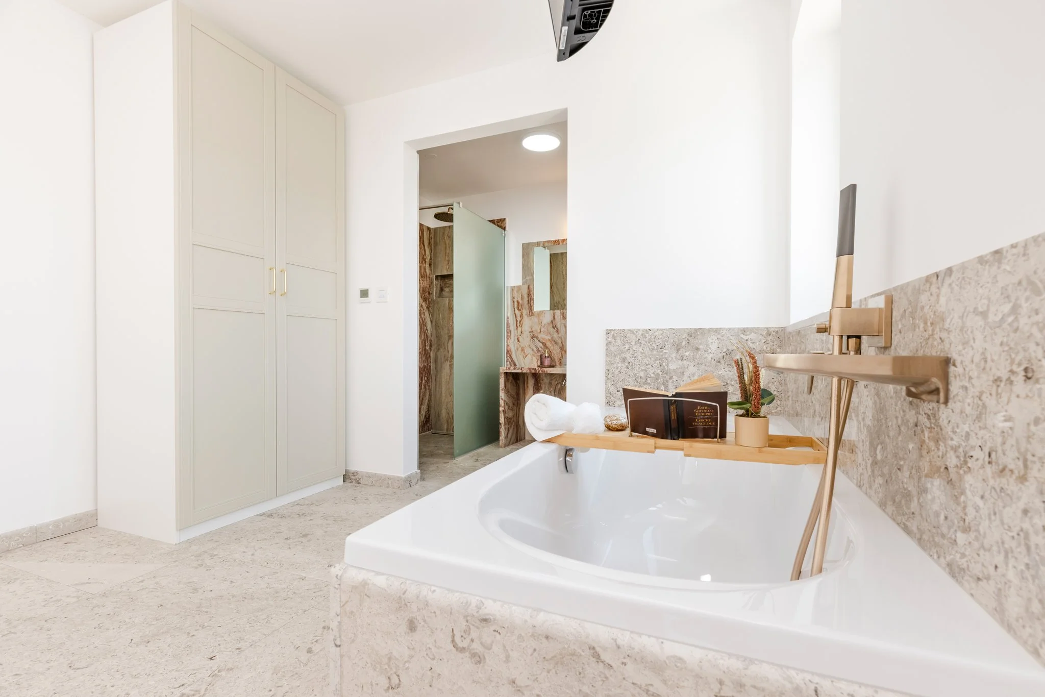 A modern bathroom featuring a bathtub with a marble surround, a wood tray holding books and decorative items, a vanity with a tabletop mirror, and a large white cabinet. The bathroom has a shower with a frosted glass door and marble wall tiles, white