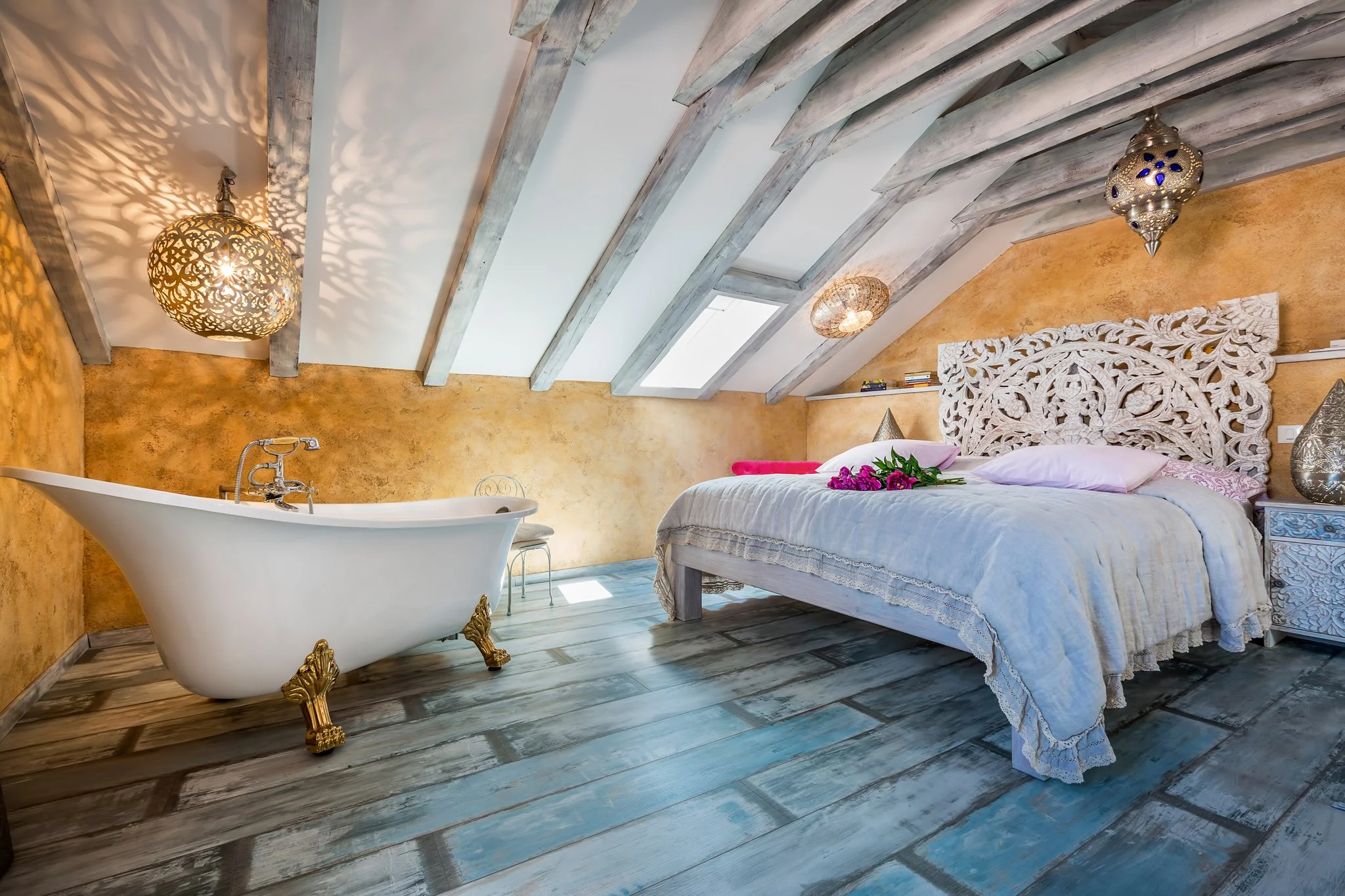A bedroom with a sloped ceiling with wooden beams and skylights. The bed has an ornate headboard, pink pillows, and a bouquet of pink flowers on top. A white freestanding bathtub with gold clawfoot legs is situated near the bed.
