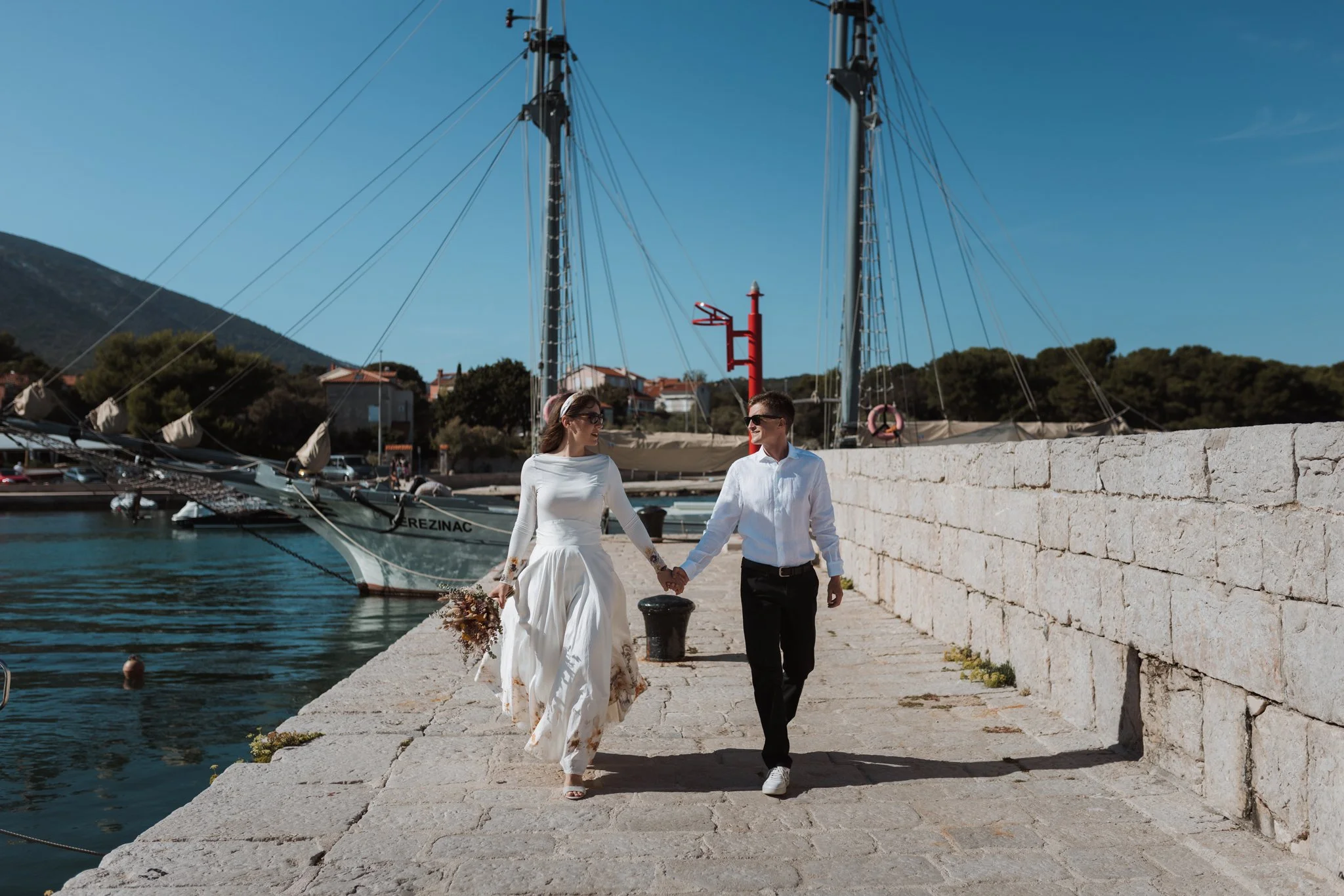 A bride and groom walk hand in hand along a stone pier at a marina, with sailboats docked nearby and a mountain in the background on a sunny day.
