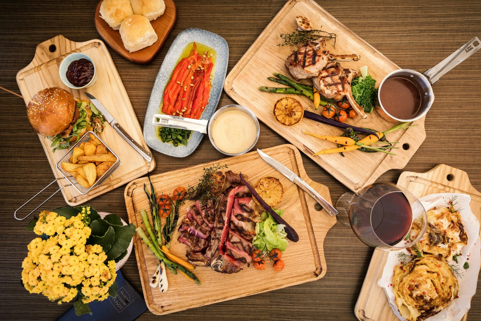 A top-down view of a variety of food dishes on wooden serving boards, including grilled meats, roasted vegetables, bread, potato wedges, a burger, and a glass of red wine, with a small pot of dipping sauce and a flower arrangement.