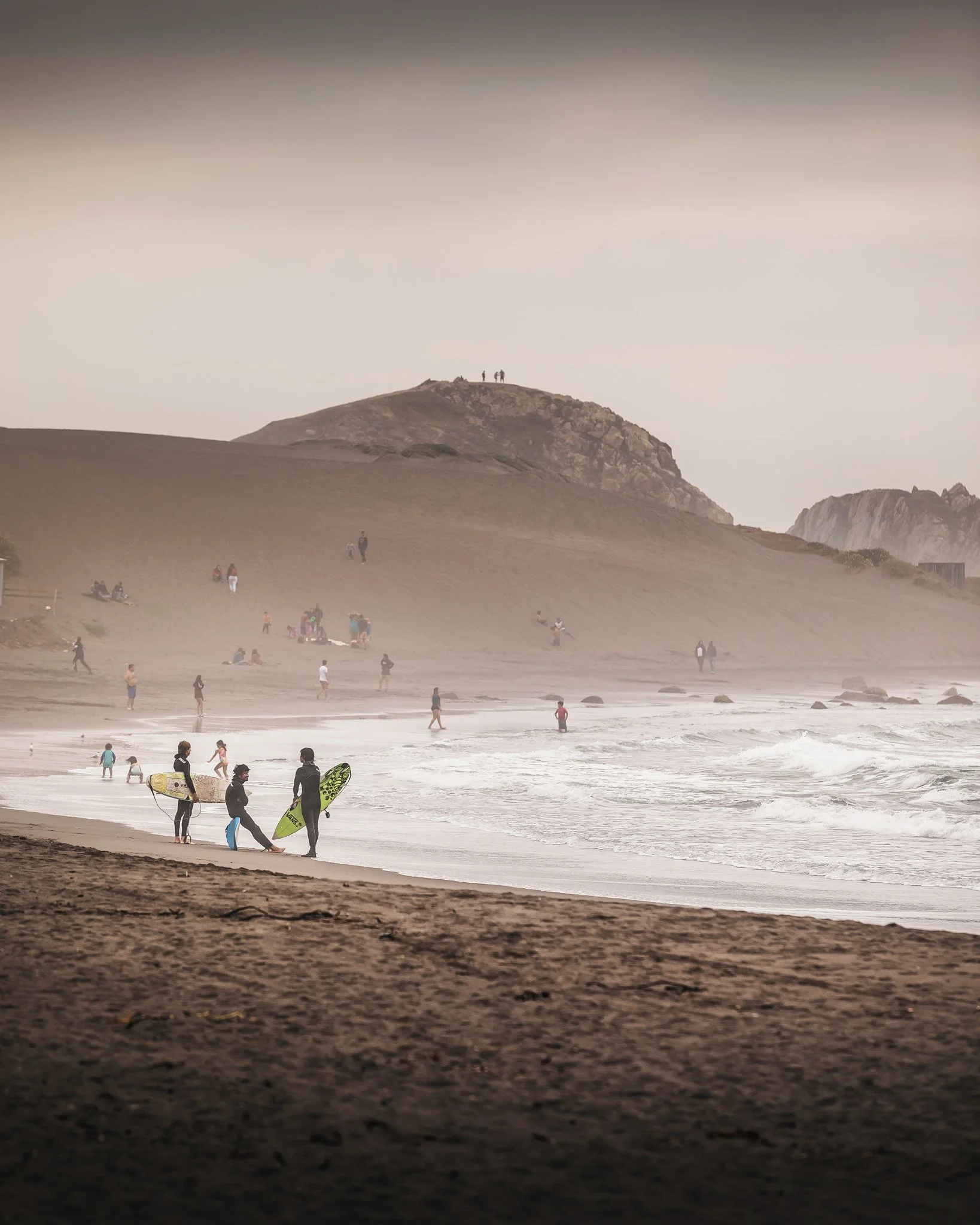 A sandy beach with people walking and engaging in activities, with rocky hills in the background and a cloudy sky overhead.