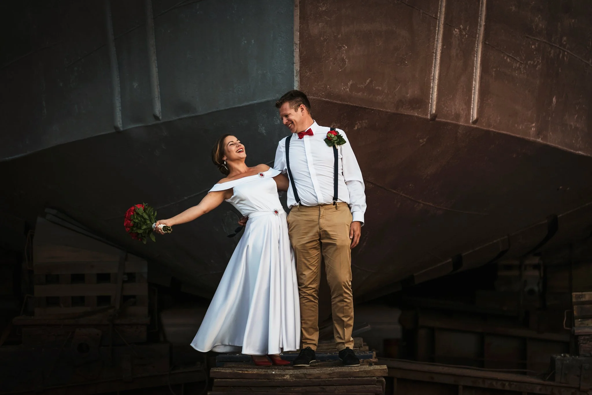 A man and woman dressed in wedding attire standing on a wooden platform underneath a large, dark metallic structure, smiling and looking at each other. The woman is holding a bouquet of red roses, and the man has a red bow tie, red boutonniere, and s