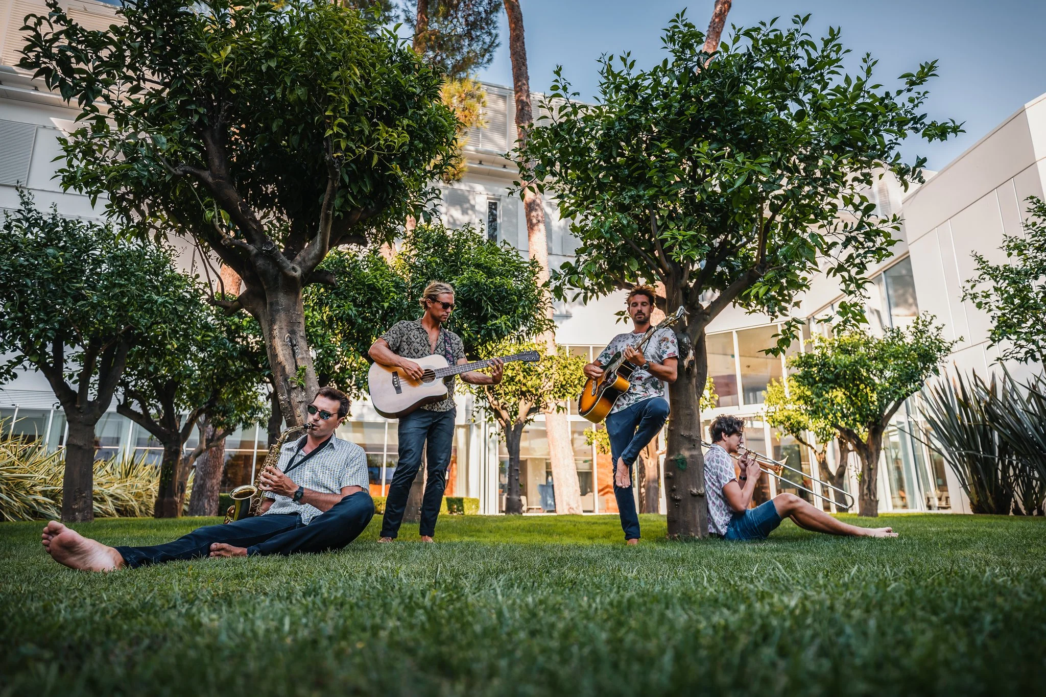 A group of five musicians performing outdoors in a modern courtyard surrounded by trees and buildings. They are playing various instruments, including guitars, saxophone, and trombone. One musician is sitting barefoot on the grass, while others are s