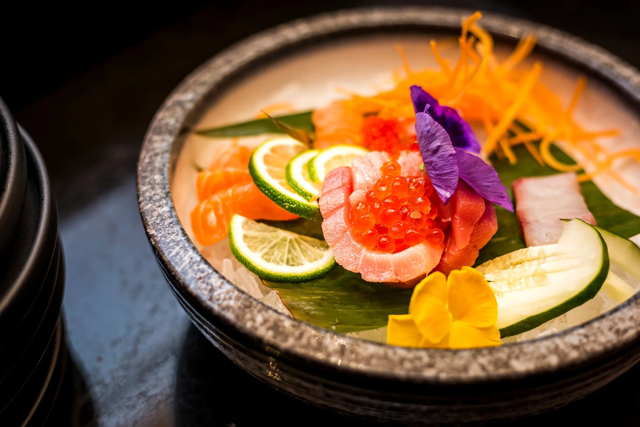 Assorted sashimi and sushi garnished with edible flowers, lemon slices, and herbs on a stone plate.