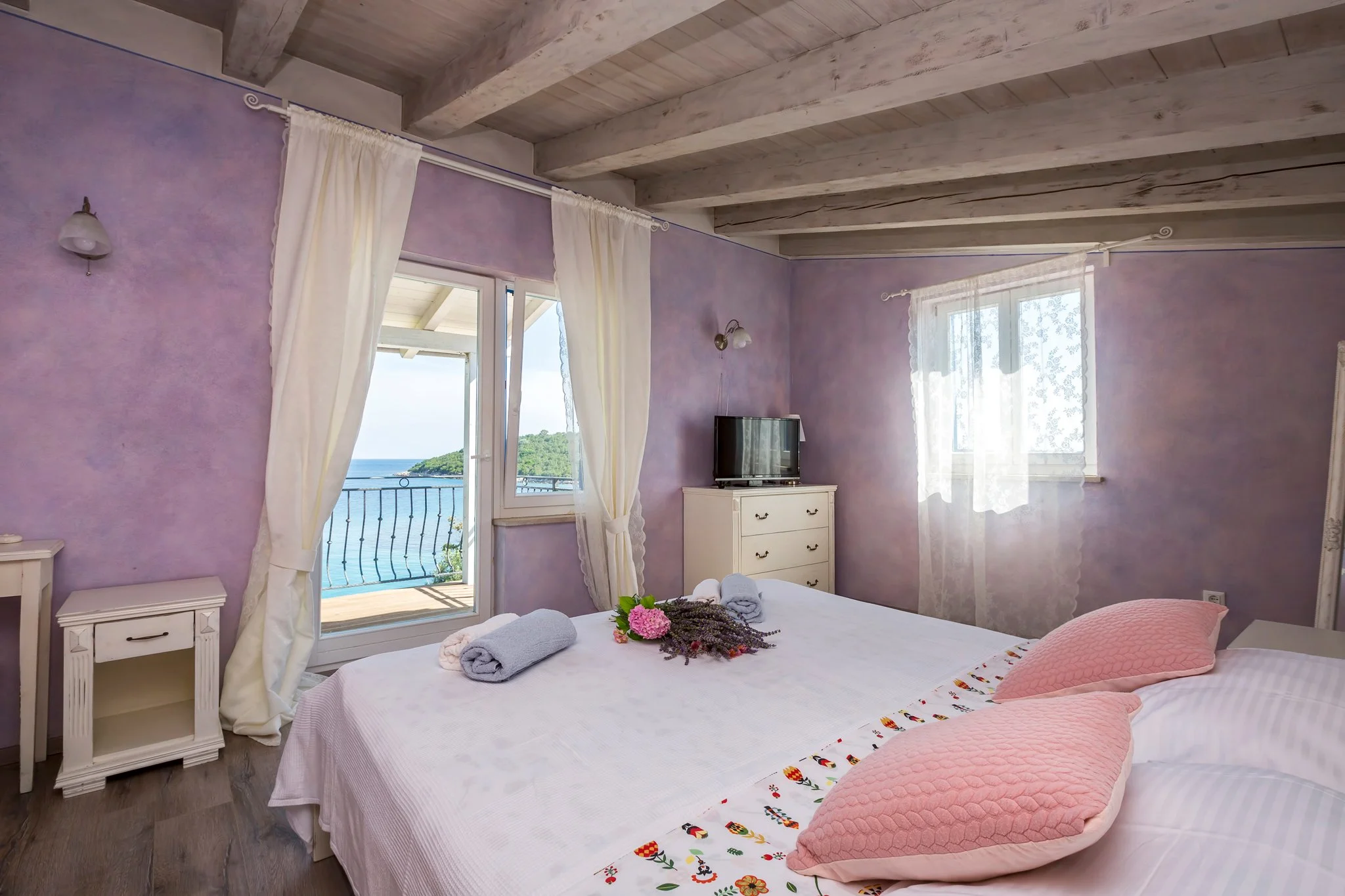 A bedroom with purple walls and white curtains. The bed has pink quilted pillows and a bedspread with embroidered details, with towels and flowers on top. There is a small white nightstand and a dresser with a TV. Large windows with views of a balcon