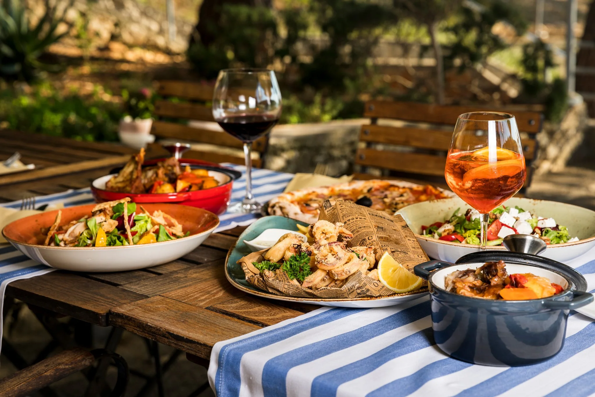 Outdoor dining table with various plates of food, glasses of red and rose wine, on a striped blue and white tablecloth, surrounded by wooden benches and greenery.
