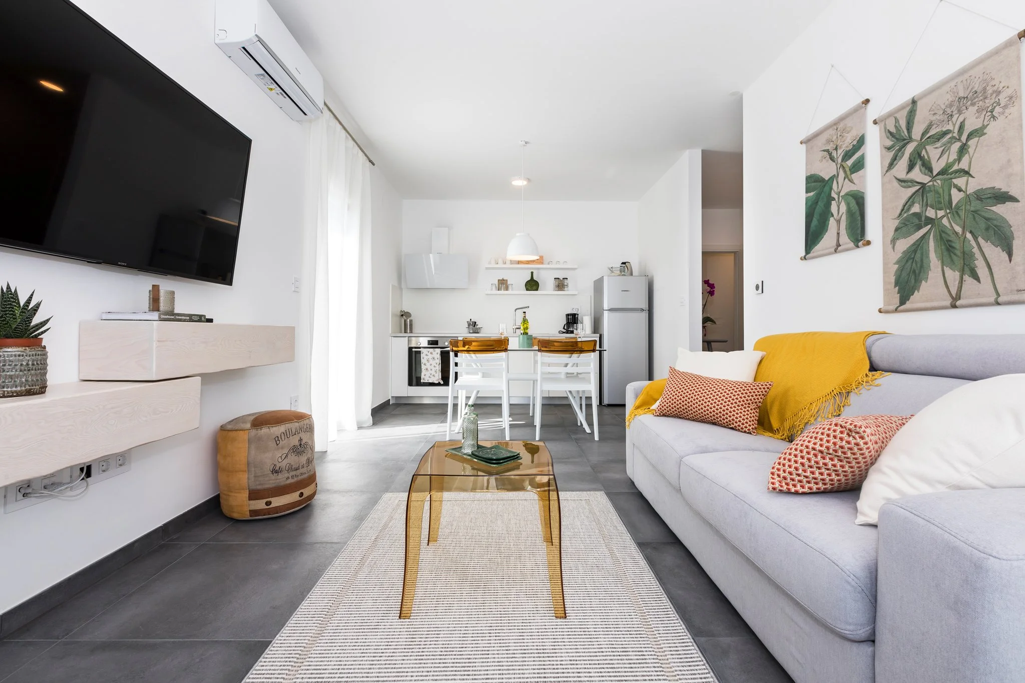Living room with white walls, gray tile floor, gray sofa with orange cushions, yellow throw blanket, glass coffee table with magazines, wall-mounted TV, air conditioner, wall art of botanical prints, open kitchen with white cabinets and dining table,