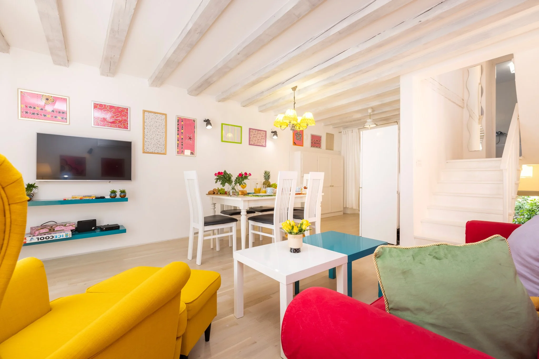 A bright living room with white walls and a sloped wooden ceiling. There is a yellow armchair and a red sofa with green and purple cushions. A white table with a flower arrangement and drinks is in the center. The wall features framed colorful abstra