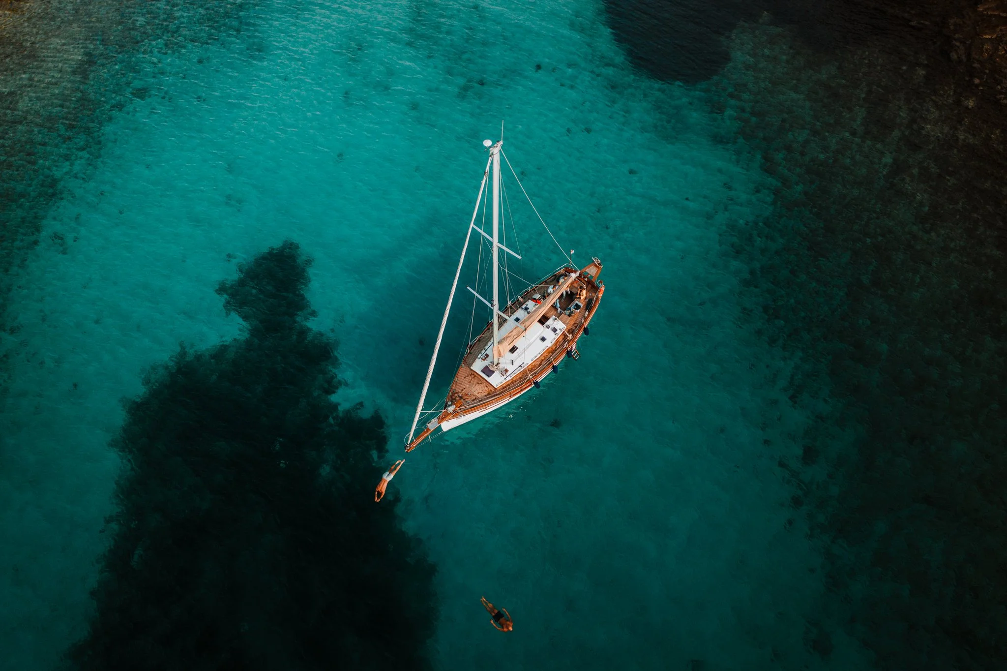 An aerial view of a sailboat floating on clear turquoise water surrounded by dark underwater land formations.