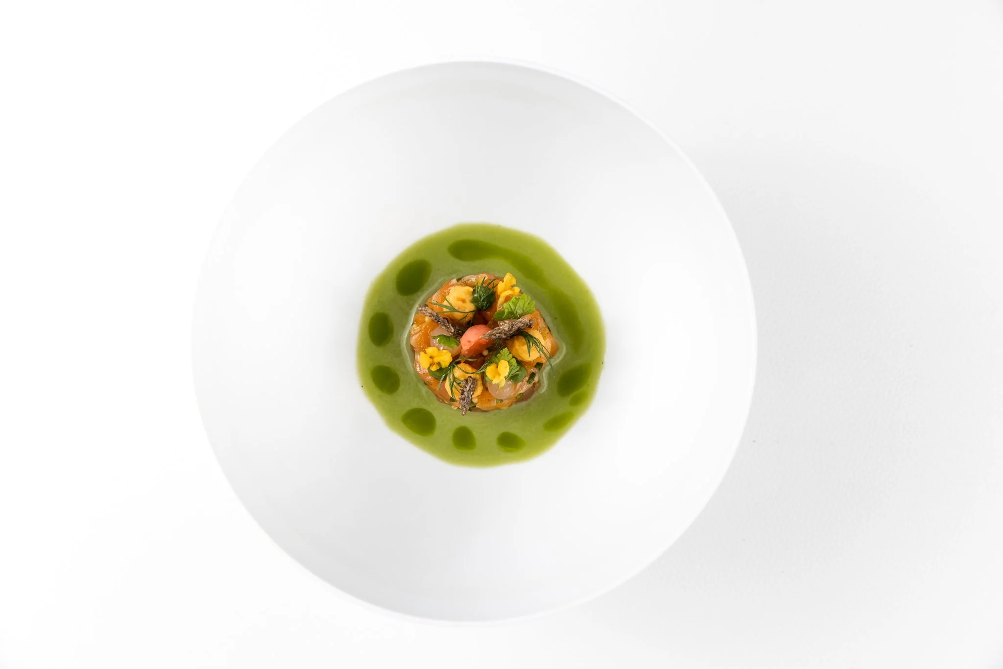 A gourmet dish with a round stack of seafood garnished with herbs and edible flowers, surrounded by vibrant green sauce on a white plate.
