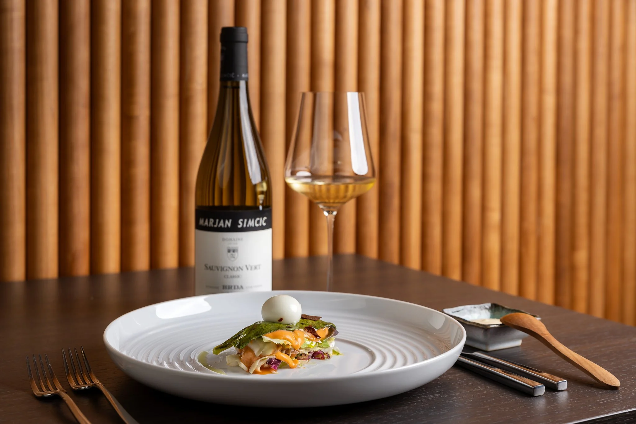 A wine bottle, a wine glass filled with white wine, and a dessert on a white plate with an artistic presentation, all placed on a dark wooden table against a background of vertical wooden slats.