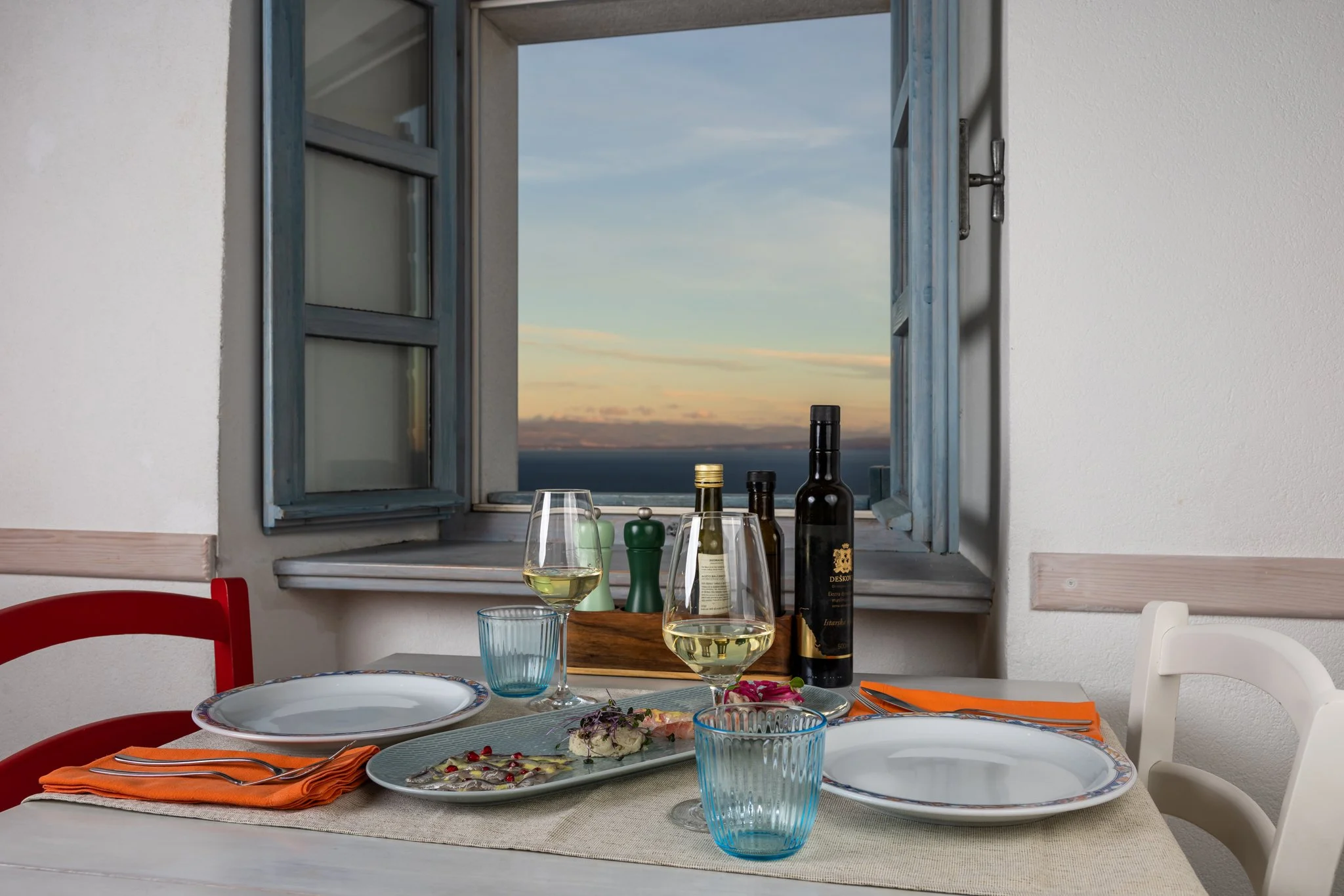 A dining table set with plates, glasses of white wine, and cutlery, with a window showing a scenic view of a partly cloudy sky and a distant horizon.