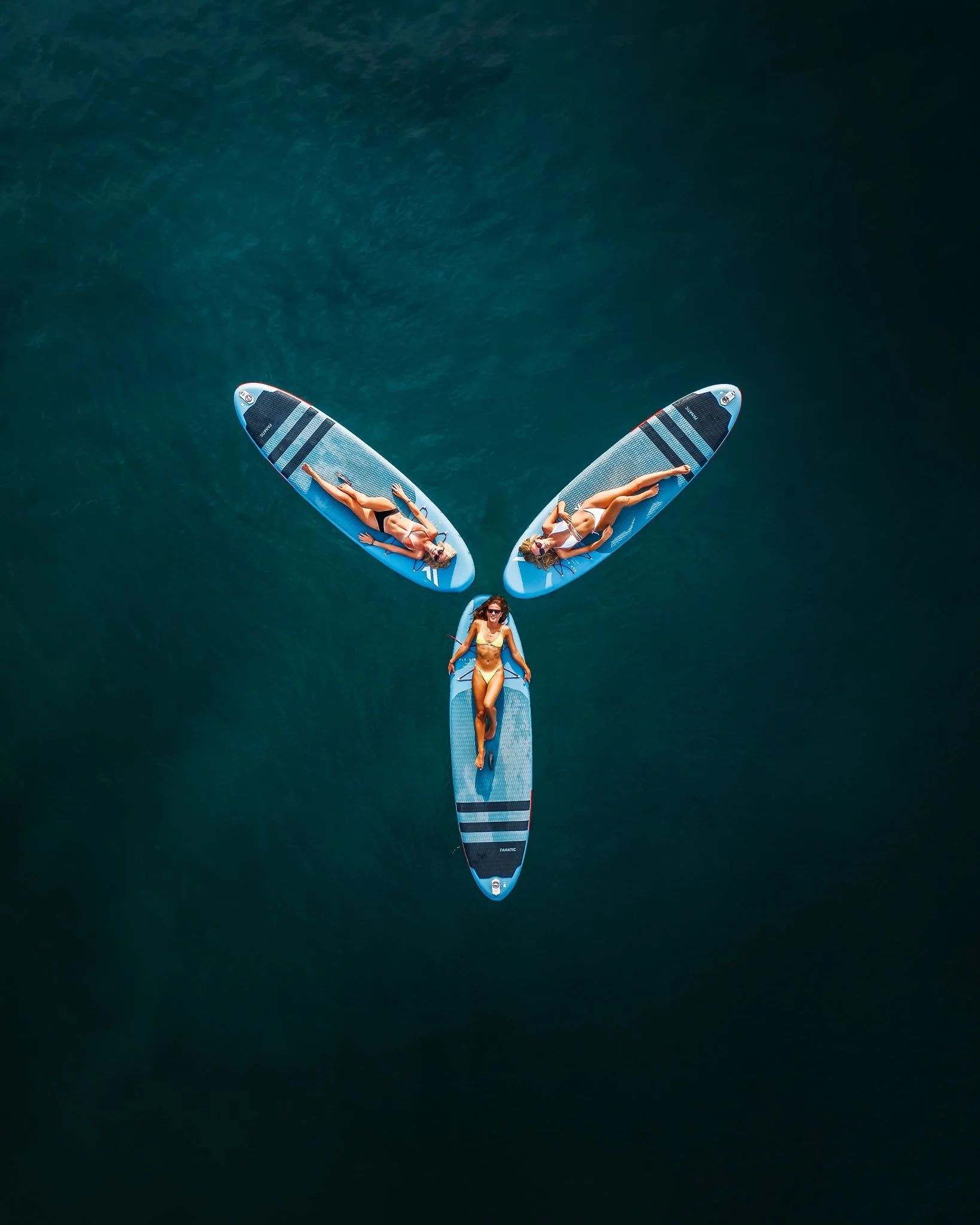 Two women lying on paddleboards in the water, viewed from above, with a third woman standing on the edge of one paddleboard.