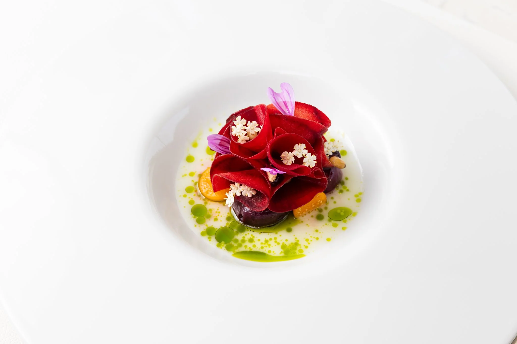 Elegant plated dish with red vegetable or fruit, edible flowers, green sauce dots, and a light drizzle on a white plate
