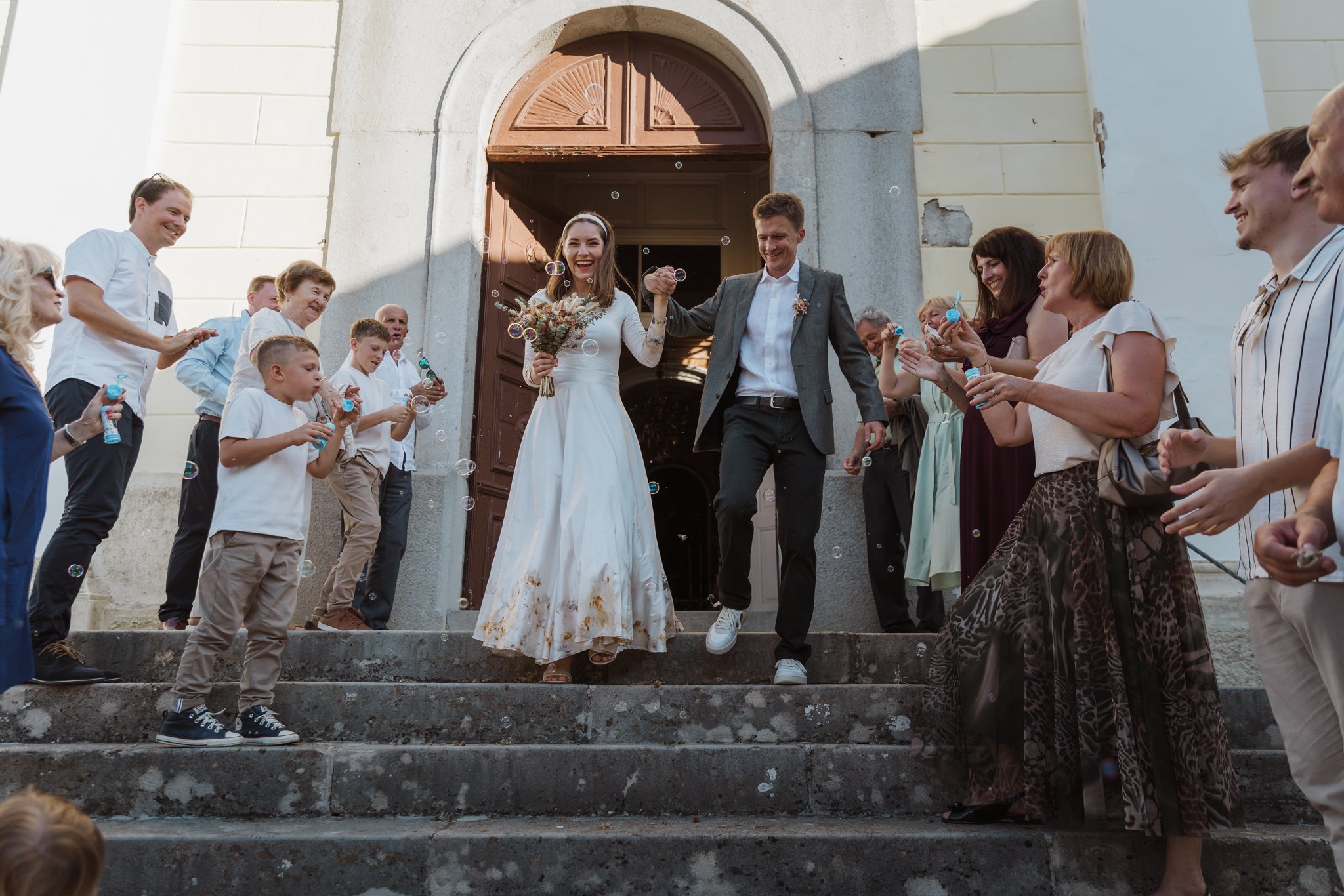 A bride and groom smiling and holding hands as they exit a building, surrounded by smiling guests blowing bubbles and clapping, on the steps of a church or cathedral.
