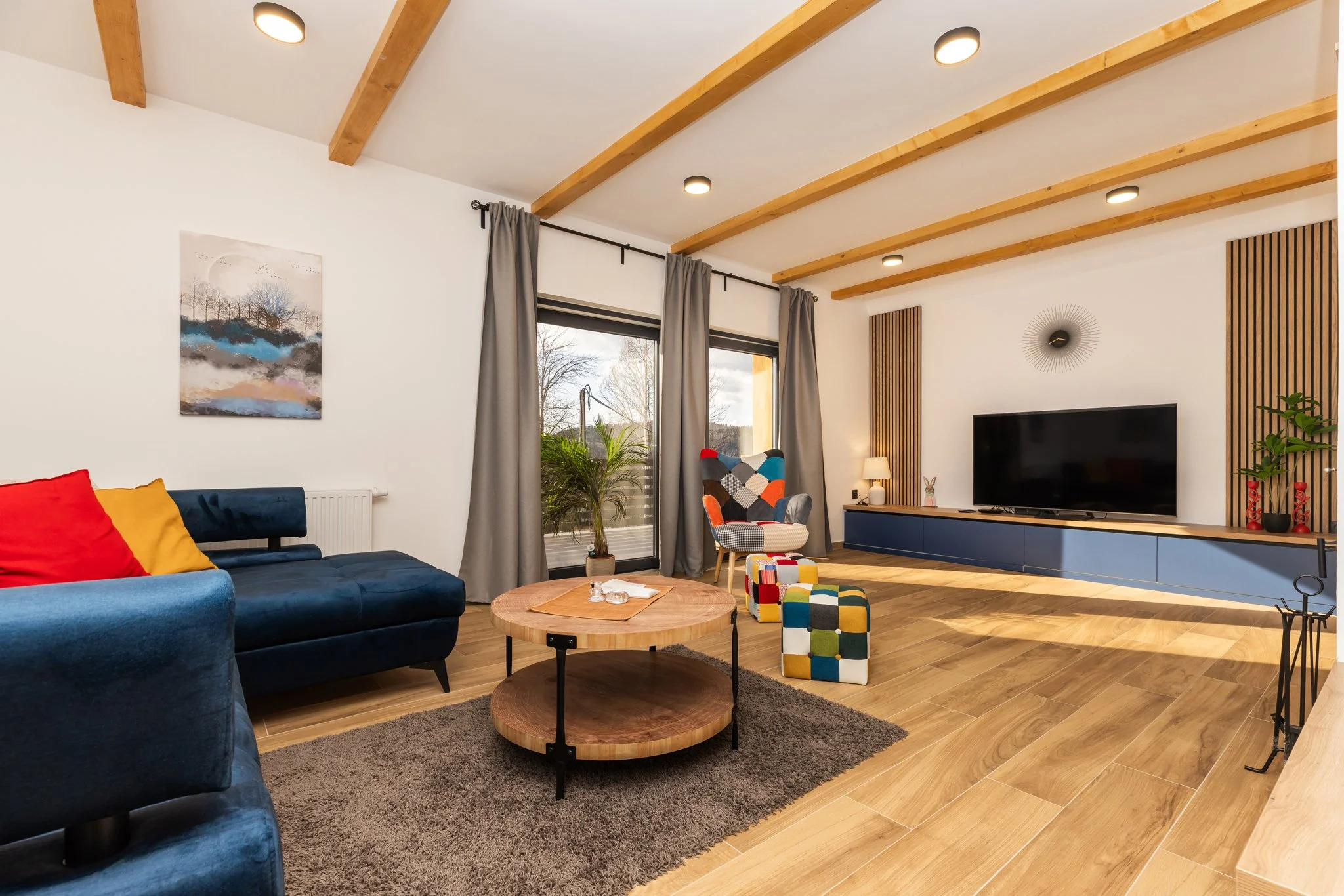 A modern living room with a wooden beamed ceiling, white walls, and hardwood flooring. It features a blue velvet sofa with colorful cushions, a round wooden coffee table, a patterned armchair, a large window with gray curtains, and a flat-screen TV o