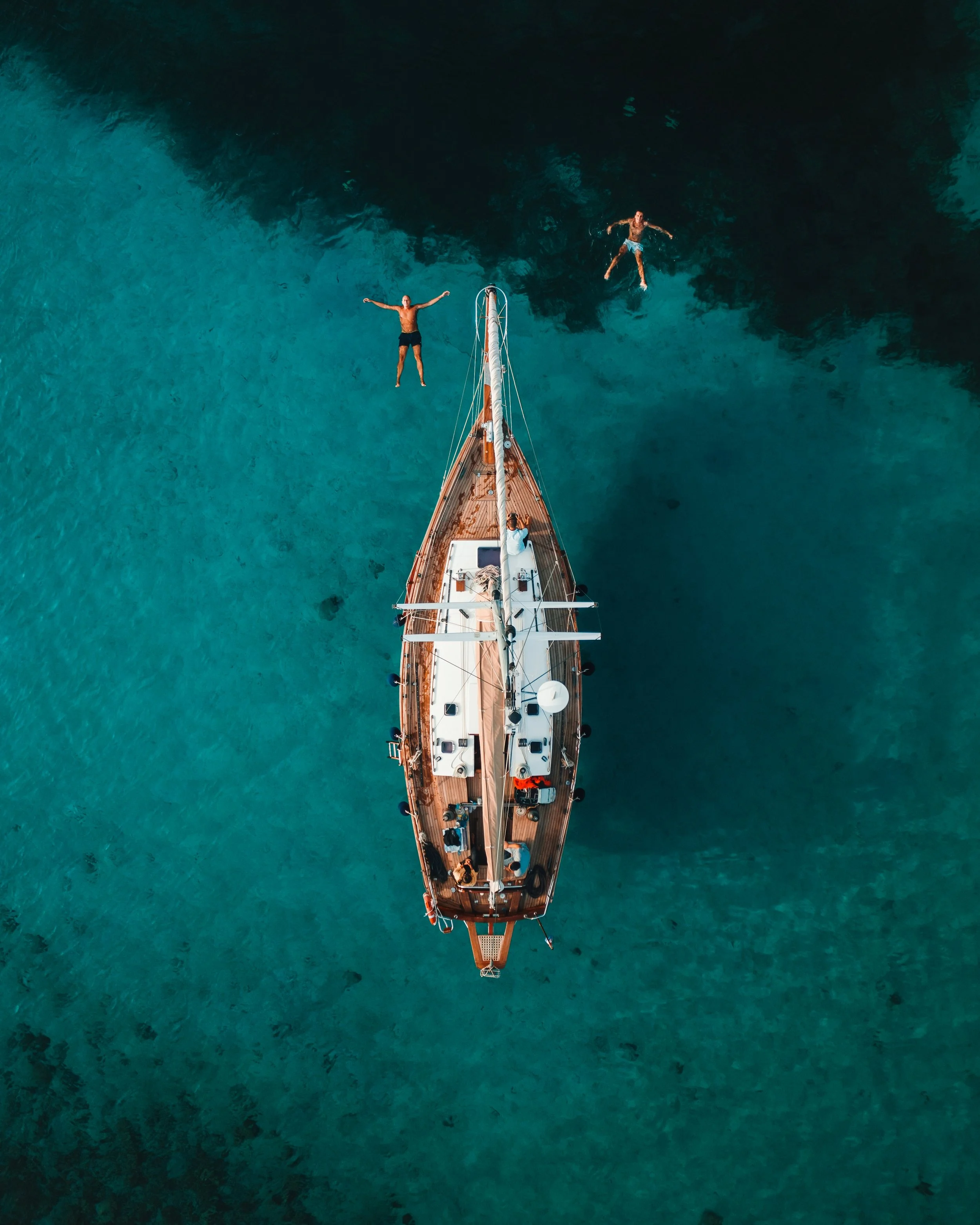Top-down view of a sailboat with four people on board and two people swimming in the turquoise water.